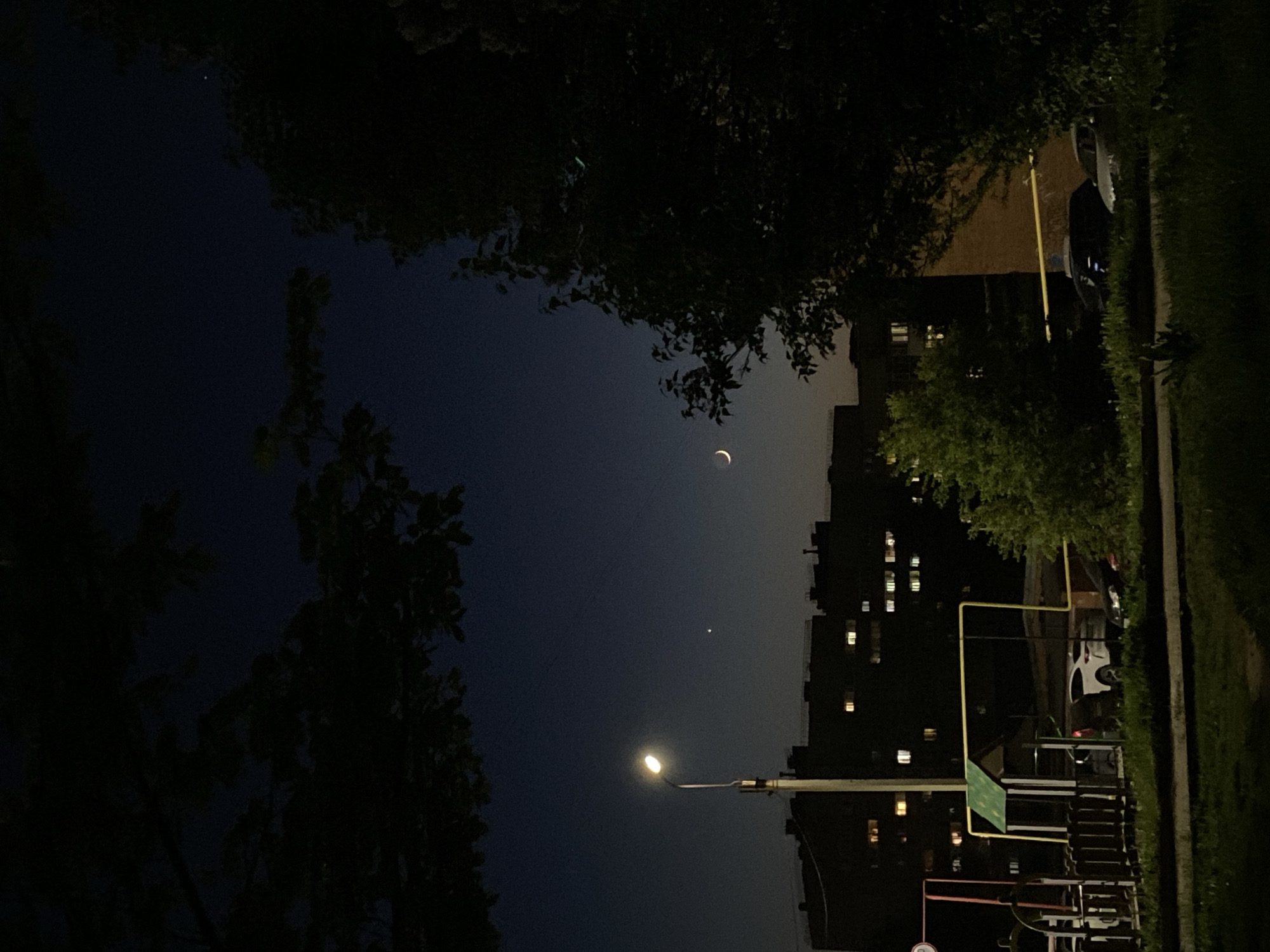 The image depicts a night scene in Votkinsk, Russia. A crescent moon and a single visible planet or star are prominent in the dark sky. Trees frame the upper part of the scene. A multi-story building with illuminated windows is visible on the right side. In the foreground, a playground structure and a street lamp are present. Parked vehicles are also discernible.