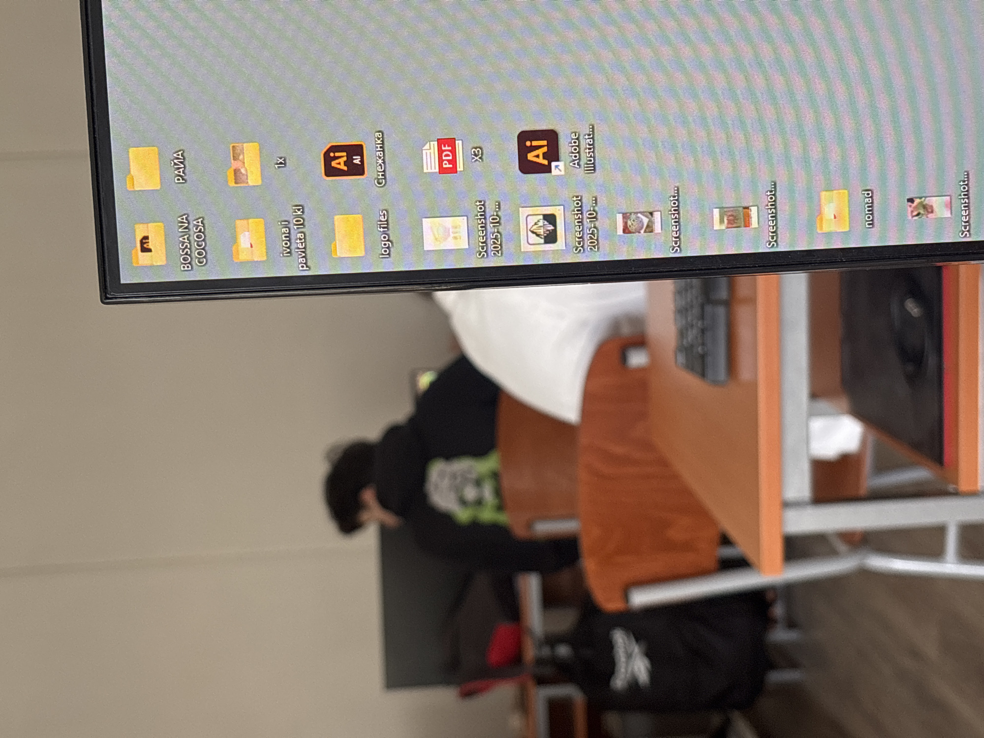 The image, taken in Troyan, Bulgaria, depicts an indoor scene with a computer monitor displaying a cluttered desktop, a person seated at a desk, and a keyboard. The monitor is positioned vertically (the image itself is rotated 90 degrees clockwise), showing numerous files and folders.
Visible text on the monitor includes folder names such as "BOSSA NA COCOSA", "РАЙА" (Raya), "ivona i pavleta 10 kl" (Ivona and Pavleta 10th grade), "logo files", "Снежанка" (Snezhanka), and "nomad". File icons and names include several Adobe Illustrator (Ai) files, a PDF document, "1x", "X3", and multiple "Screenshot" files, some dated "2025-10-...".
A person, visible from the back, wears a black hoodie and a black beanie, seated in a wooden chair at a light-colored wooden desk with a metal frame. A keyboard is visible on the desk, partially obscured. The background shows plain white walls. The setting appears to be a classroom, computer lab, or office. The person is engaged in an activity on the computer, though the specific task is unclear. The Cyrillic text on the monitor reinforces the Bulgarian location context.