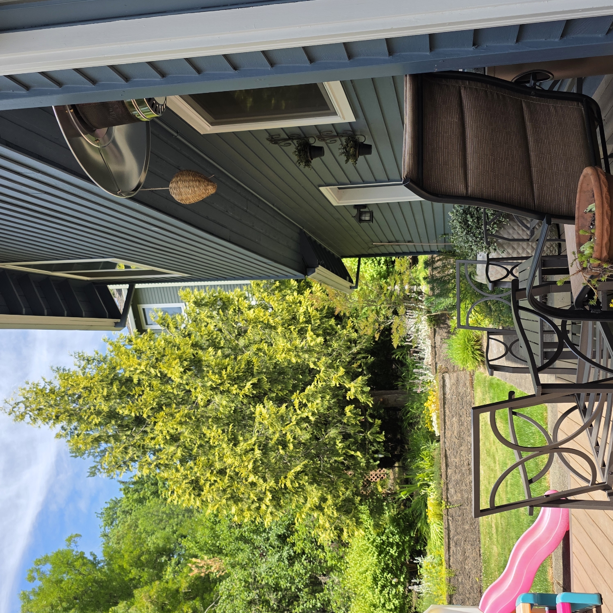 This media file shows an outdoor patio area in Portland, Oregon. The scene features a covered patio with a large, vibrant green tree in the background. The patio is furnished with outdoor seating, including a brown upholstered chair and a black metal table and chairs. A pink slide is visible on the wooden deck.
The house siding is a dark teal color, and a patio heater is mounted overhead. There are also decorative items, such as small planters with greenery hanging from the eaves and a woven basket ornament. The sun is shining, suggesting it is daytime and likely pleasant weather. The overall atmosphere is relaxed and suitable for outdoor leisure. No people are visible in the image. There is no visible text.