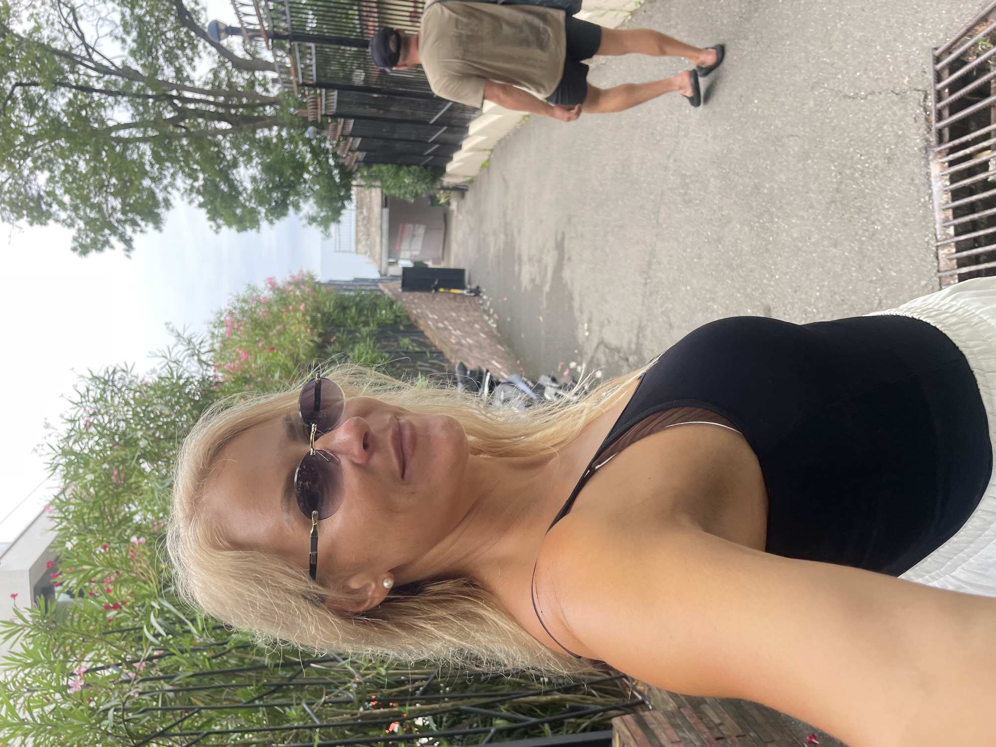 A woman with blonde hair and sunglasses takes a selfie on a street in Sochi, Russia. She is wearing a black tank top and white pants. Behind her, a man in a tan t-shirt and black shorts walks away from the camera. The street is paved and lined with a fence on the right and a brick wall with plants on the left. In the distance, a body of water can be seen.
