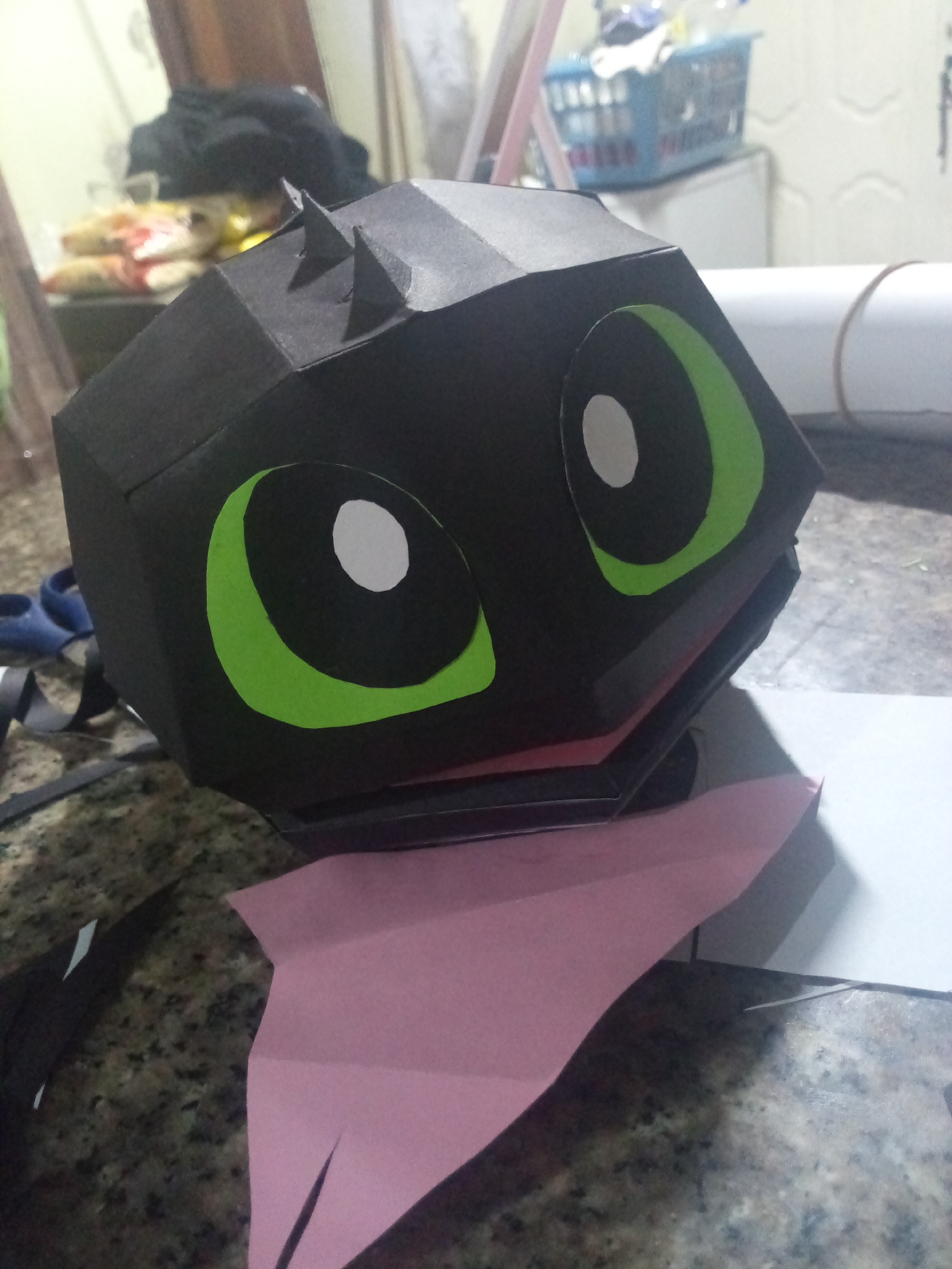 On a speckled brown countertop, a partially-constructed craft of Toothless from the movie "How to Train Your Dragon" sits. The head is a black, geometric shape with applied green and white eyes and small spikes on top. Below the head is a red section and a pink tongue or bib made of paper. In the background, there's a cluttered scene with objects including a blue basket, rolled paper, and bags of assorted items on a white surface. The image appears to be taken in an indoor setting, likely a home or craft area. There is no information indicating the city of El Valle del Espíritu Santo, Venezuela.