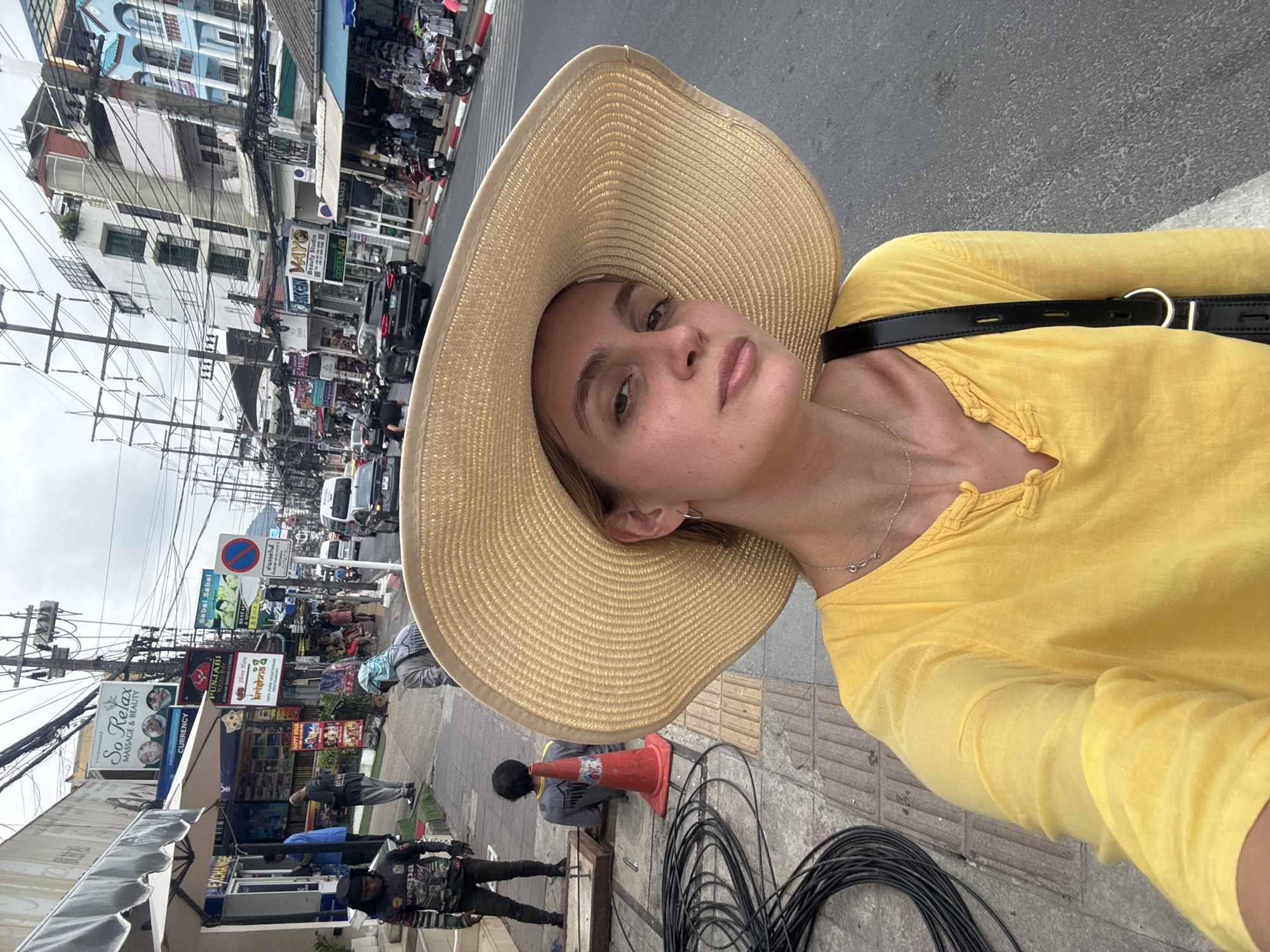 The image captures a scene on a street in Patong, Thailand. A person wearing a large, tan sun hat and a yellow shirt is taking a selfie. A black bag strap is visible over the person's shoulder. The background reveals a bustling street with various shops, signs, and overhead wires. There's a no parking sign in Thai and English, along with signs for businesses such as "So Relax Massage & Beauty" and "Punjabi King of Indian Cuisine." A man is kneeling near a traffic cone, possibly working on cables lying on the sidewalk. Other people are walking along the street. Buildings of varying heights and colors line the street, and there's moderate traffic.