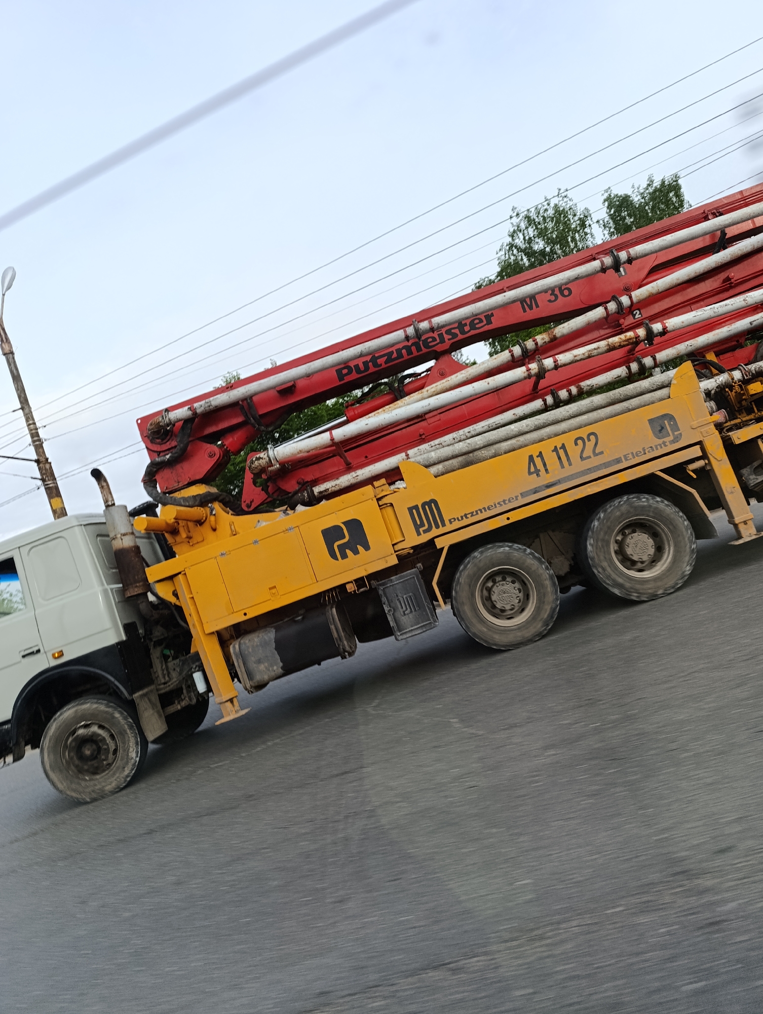 The image shows a Putzmeister M36 concrete pump truck in motion on an asphalt road in an urban area. The truck has a white cab and a yellow chassis, with a red boom assembly. The company logo is visible on the side of the chassis, and the number "41 11 22" is displayed. The background includes trees, power lines, and a street lamp. The sky appears overcast.