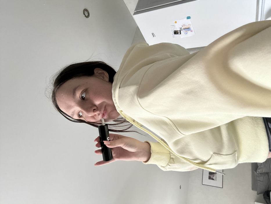 The scene depicts an indoor environment, likely a residential kitchen or living area, in the city of Chaikovsky, Russia. A female individual, with dark hair, is positioned in the foreground, facing slightly away from the camera. She is wearing a light yellow hooded sweatshirt. Her right hand holds a black, cylindrical electronic device, likely a vape pen, with its mouthpiece at her lips. A small red indicator light is visible on the side of the device. The woman's head is tilted slightly upward, and she is looking towards her upper right. In the background to the right, a white Beko brand refrigerator is visible, adorned with several papers or magnets. To the left, a portion of a dark grey sofa and a framed picture on a white wall are discernible. The ceiling is white, featuring a recessed circular light fixture.
