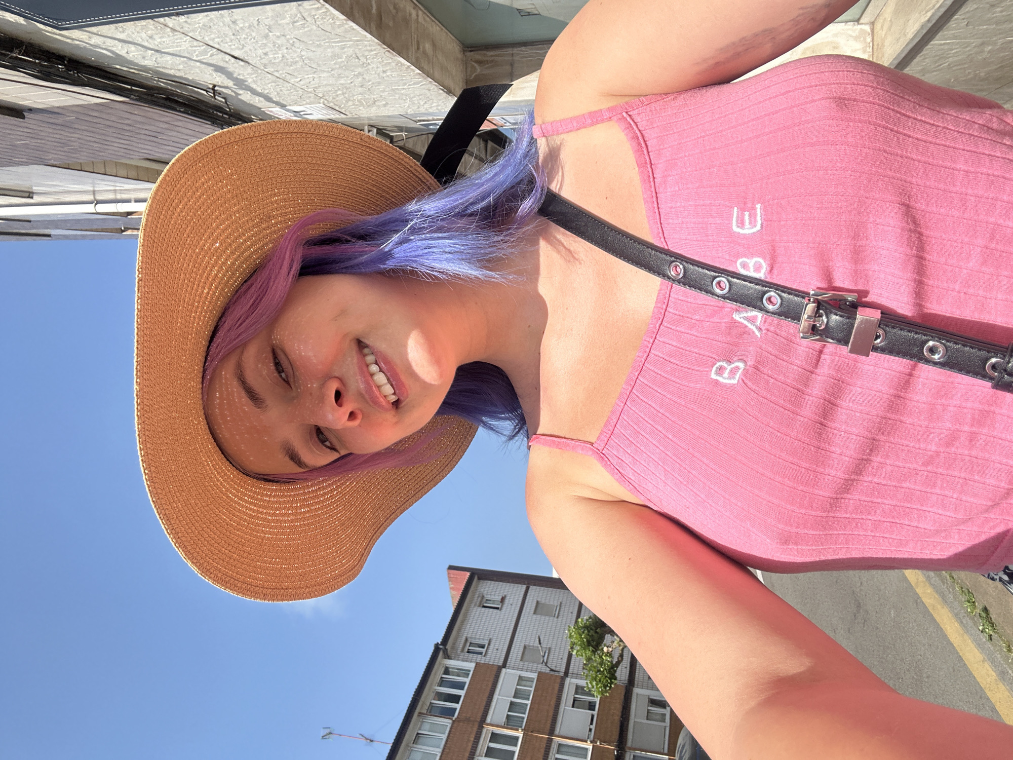 A young woman, photographed from a low angle, is the central subject. She wears a light-brown sun hat and a pink tank top with the word "BABE" embroidered on it. A black purse strap is slung across her torso. Her hair is dyed purple. She is smiling at the camera. The background includes a clear, blue sky, a building with a tiled exterior and multiple windows, and a street with a yellow line. The location appears to be Gijón/Xixón, Spain, due to the style of architecture and urban setting.