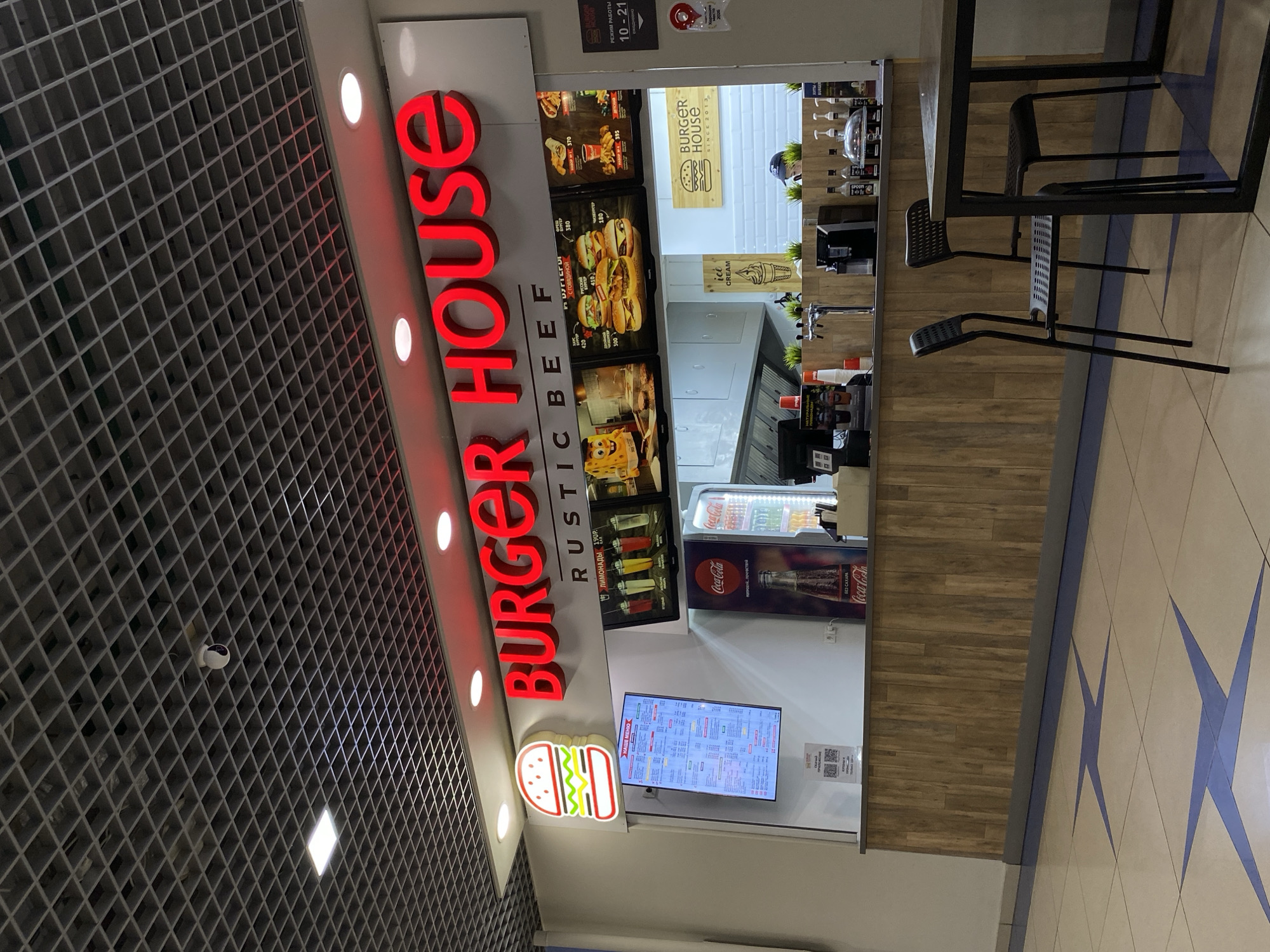 The image shows the interior of a Burger House restaurant located in Votkinsk, Russia. The restaurant features a modern, clean design with a prominent red "BURGER HOUSE" sign and images of burgers displayed on a digital menu board. The counter area has a wooden facade and a selection of drinks and condiments. Two chairs are visible in the foreground, suggesting a dining area. The overall atmosphere appears to be that of a fast-food establishment.