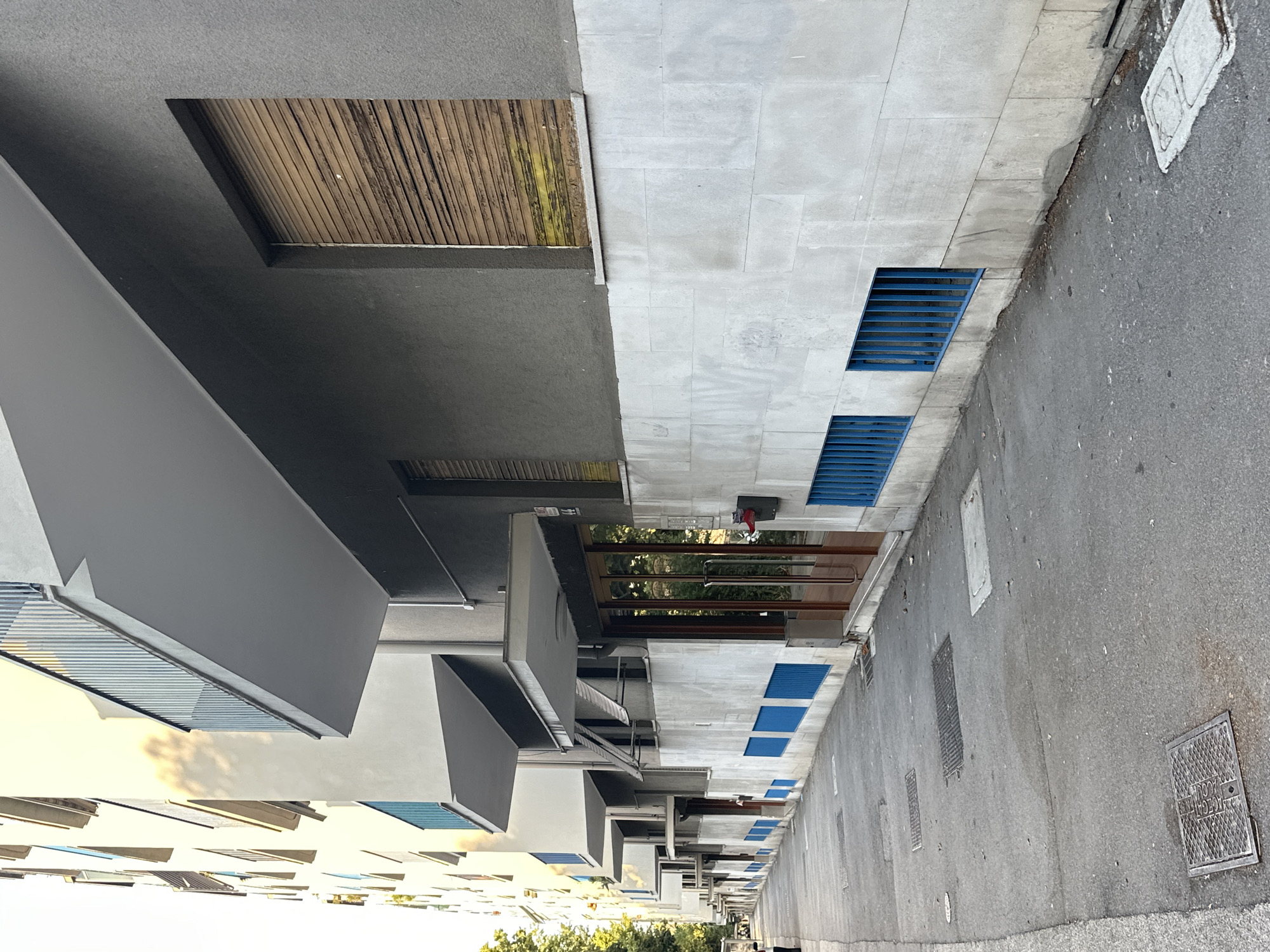 The image captures a street-level view of a modern apartment building with a repeating architectural design, likely in Trieste, Italy. The building features a grey facade with a lower section clad in light-colored stone or concrete blocks. Several windows are covered with weathered wooden roller shutters. Below these shutters, there are rectangular openings with bright blue metal grates. The building has balconies with metal railings extending from the upper floors, and under-balcony structures that cast shadows on the street. A glass-paneled entrance door with wooden framing is visible on the ground floor, along with a red fire extinguisher mounted on the wall to its left. The sidewalk is paved with asphalt and features several manhole covers and drainage grates. Trees and foliage are visible on the far left, suggesting a street lined with greenery. The overall scene is calm and static, with no people prominently visible in the immediate frame. The lighting suggests it is daytime, possibly late afternoon, given the long shadows.