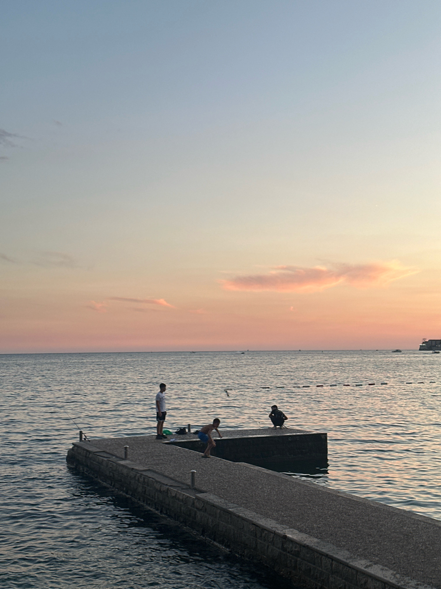 This image depicts a sunset scene at a pier in Budva, Montenegro. The sky transitions from a pale blue at the top to soft pinks and oranges near the horizon, with delicate clouds illuminated by the setting sun. The sea is calm with gentle ripples reflecting the colors of the sky.
On the pier, three individuals are visible. One person, appearing to be a young man, stands near the edge of the pier, looking out towards the sea. Another person, also a young male, is crouched down on the pier, seemingly engaged in an activity near the water's edge. A third person is seated on the edge of a higher section of the pier. A fishing rod is set up near the edge of the pier. In the background, several small boats are scattered on the water, and a buoys line marks a section of the sea. The pier itself is constructed of concrete and stone, extending into the water. A building or structure with trees is visible in the far distance on the right side of the image.