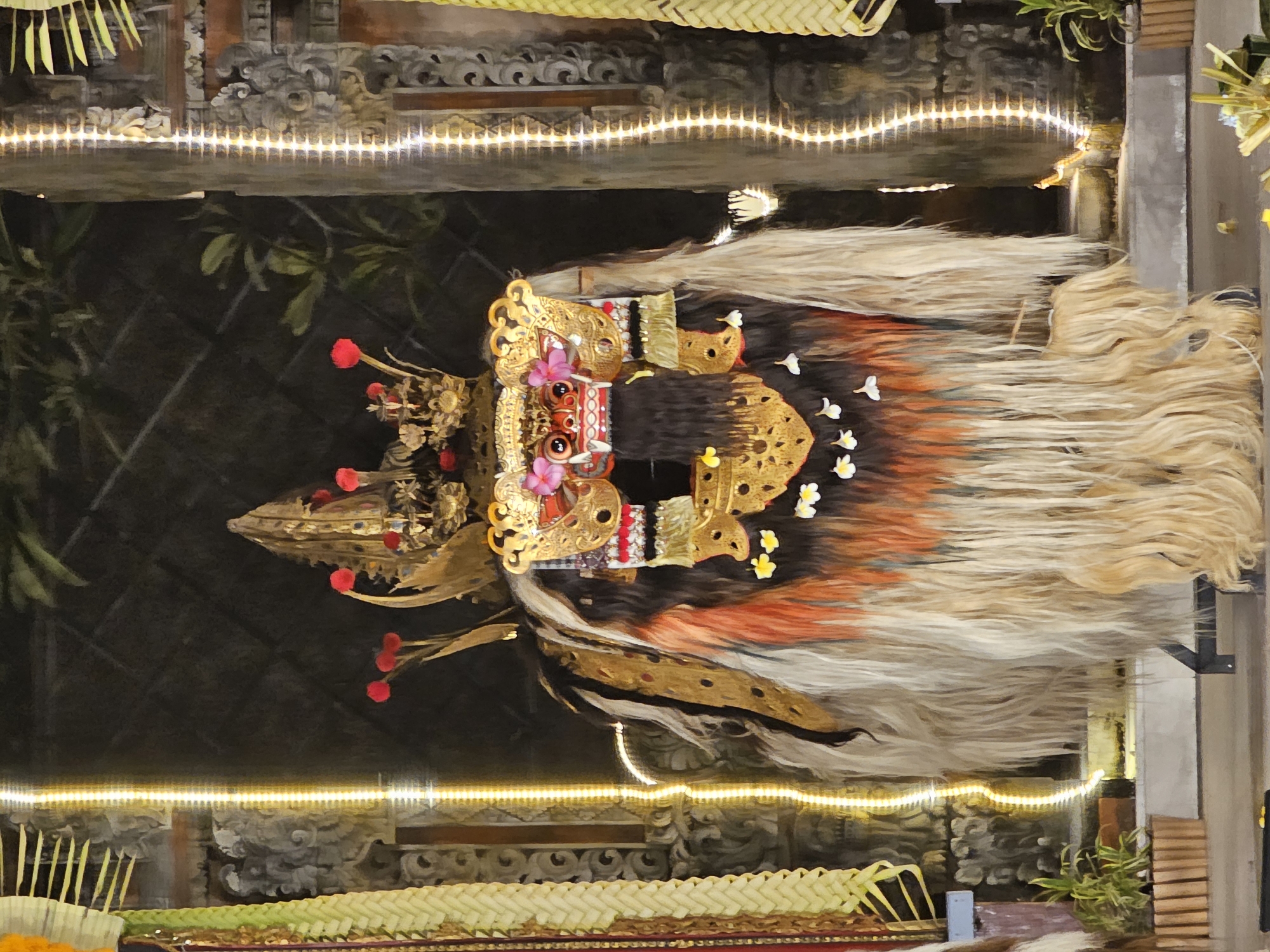 This image shows the Barong dance costume, which is used in the Denpasar area of Indonesia. The Barong is a panther-like creature and character in the mythology of Bali. The costume is adorned with gold trim and includes a mask with protruding eyes. A long mane of light-colored hair drapes down the sides. The display is set up with woven matting and candles, suggesting a ceremonial or display setting. The background features intricate stone carvings and leafy plants.