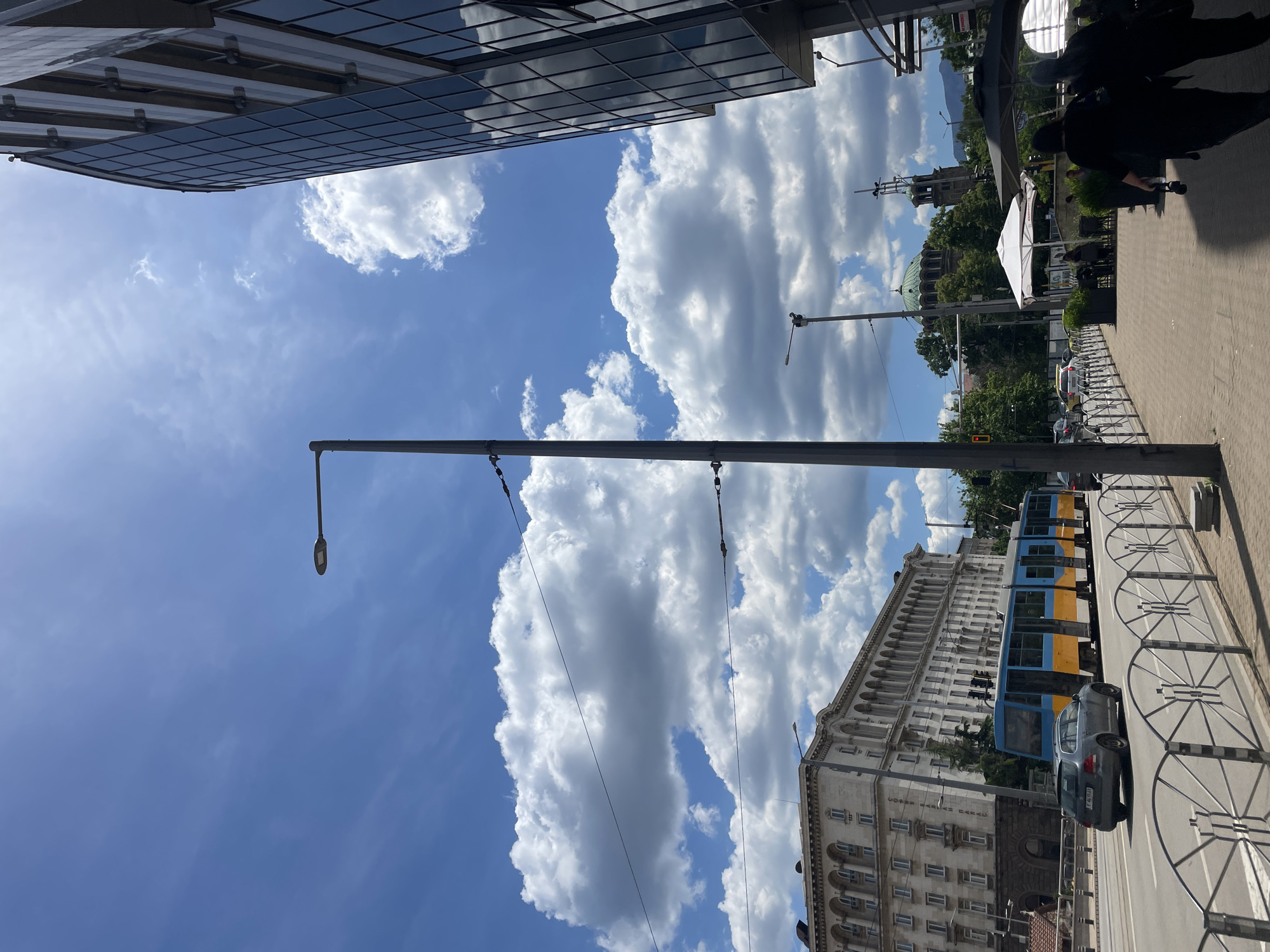 The image captures a street scene in Sofia, Bulgaria, looking upwards. A modern glass-faced building is visible on the left. Dominating the sky are puffy white clouds against a bright blue background. A tall, slender light pole stretches upwards, bisecting the view. On the right side of the frame, there's a pedestrian sidewalk, bordered by a decorative metal fence. A section of a blue and yellow tram is visible, along with a parked gray car. In the background, the Alexander Nevsky Cathedral and other buildings are seen beyond the trees and street furniture. A person is walking on the sidewalk.