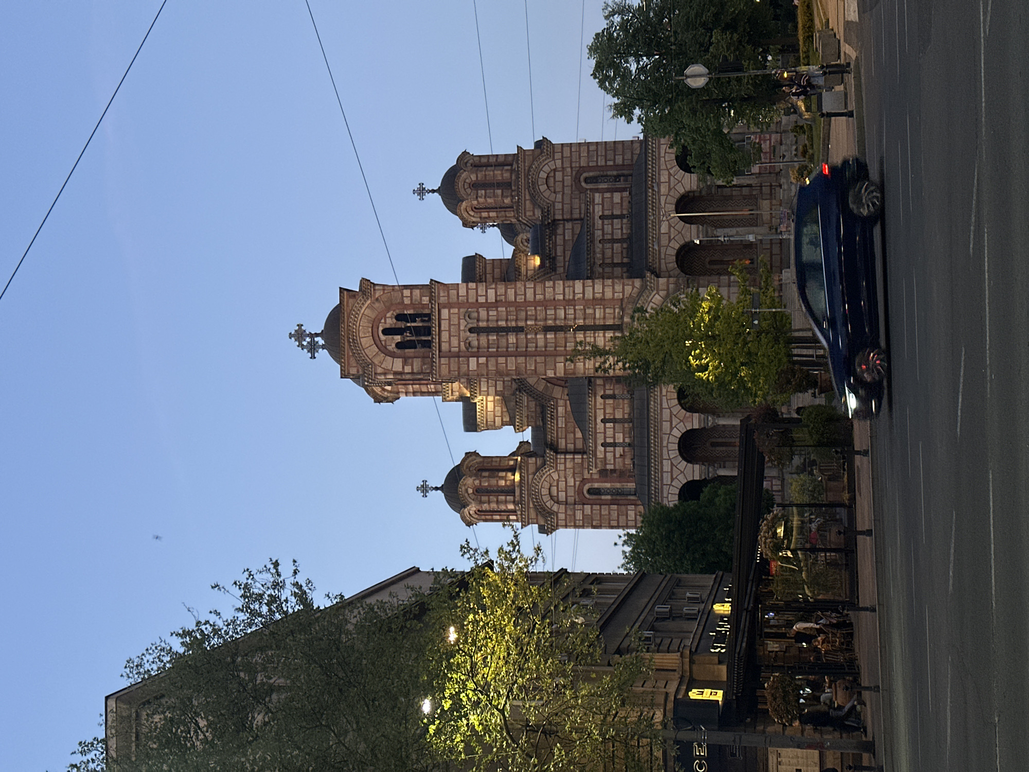 The image shows the Church of Saint Mark in Belgrade, Serbia, at dusk. The church, a large structure with multiple towers and crosses, is made of red and white stone. Power lines are visible against the sky. A blue car is driving on the road in the foreground. On the right is an outdoor restaurant with tables and chairs, some occupied by people. Trees are interspersed throughout the scene, some with lights. Another building is visible on the left.