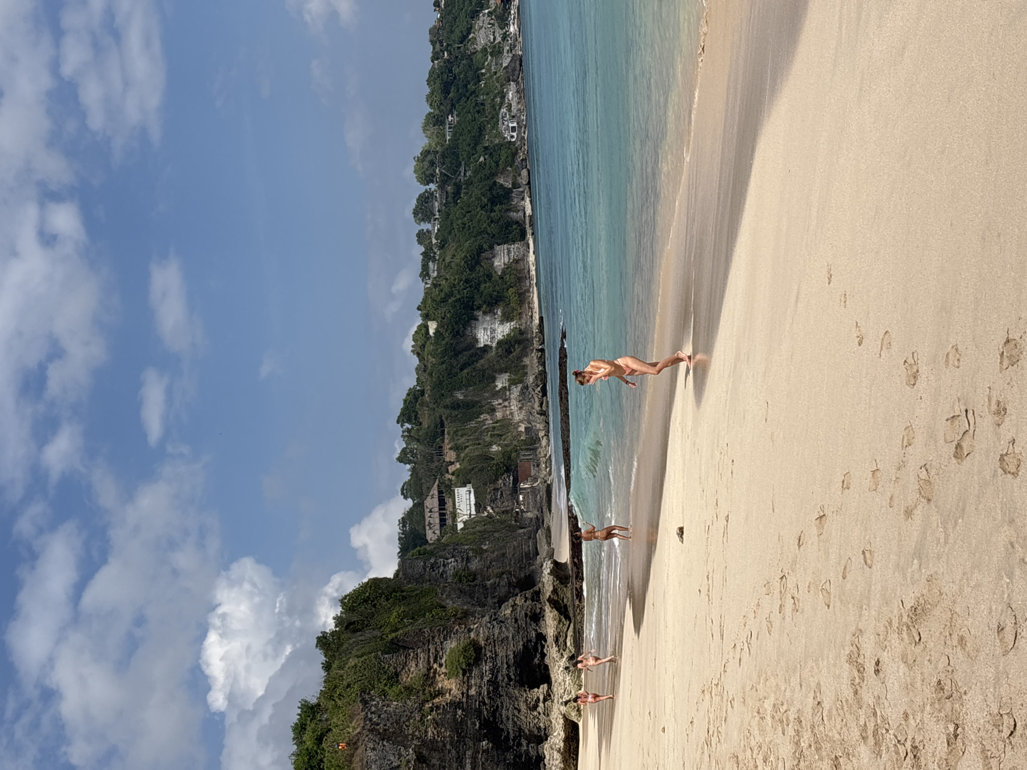 The image shows a scenic view of Bingin Beach in Kuta Selatan, Indonesia. The beach is characterized by its sandy shore, clear turquoise waters, and dramatic limestone cliffs covered in lush green vegetation. In the background, buildings and resorts are visible nestled atop the cliffs.
Several individuals are present on the beach, enjoying the sunny weather. Three women are walking along the shoreline, some wading in the shallow water. One woman is captured mid-stride, appearing to be walking towards the ocean, while another is walking away from the water's edge. A fourth person is visible further down the beach, partially obscured by the waves. The sand is marked with footprints, indicating recent activity. The sky is a vibrant blue, dotted with fluffy white clouds. The overall atmosphere is one of relaxation and enjoyment of a tropical beach setting.