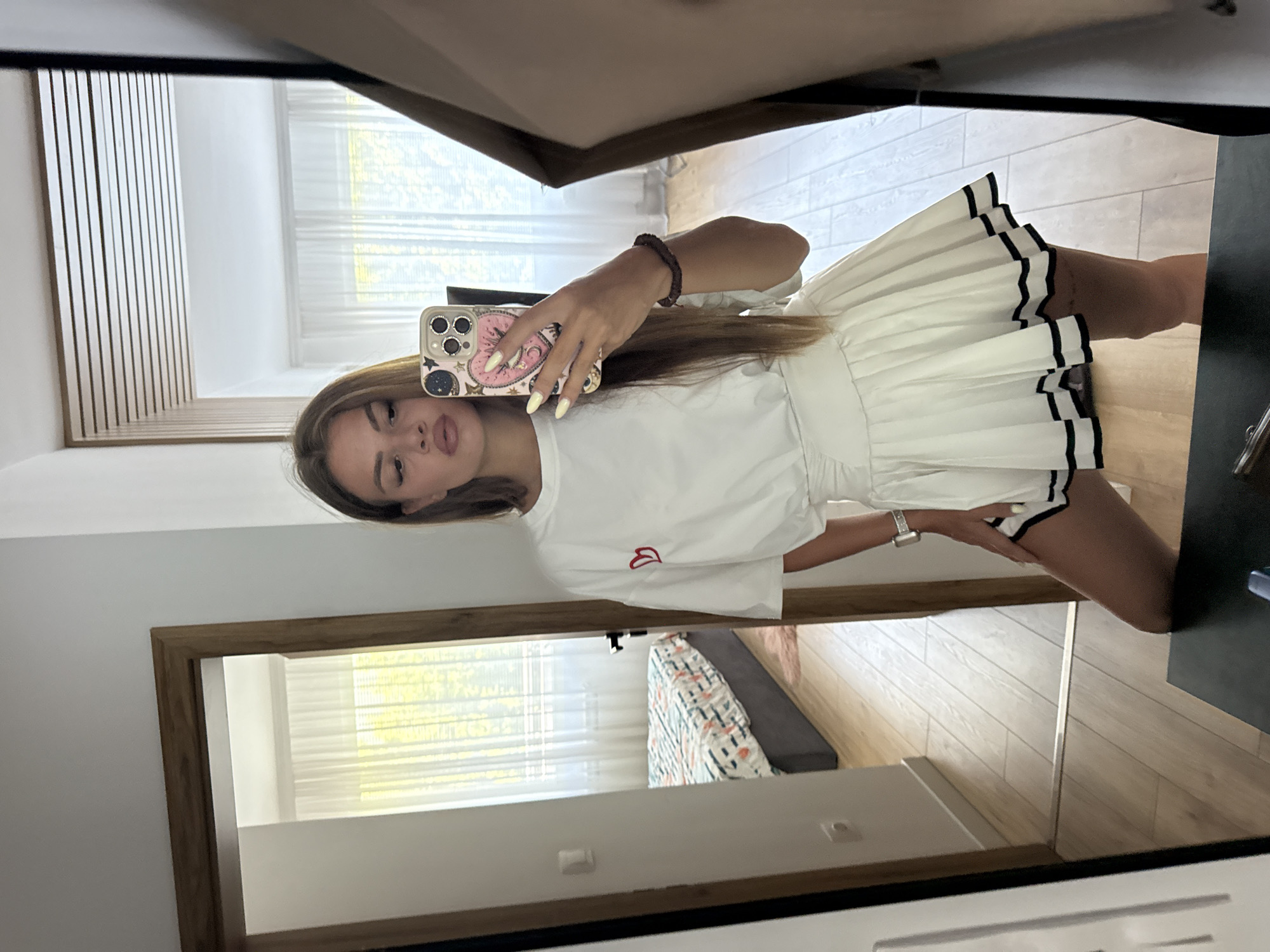 A young woman is taking a mirror selfie in what appears to be a bedroom. She is wearing a white t-shirt with a small red heart design on the left chest and a white pleated skirt with black trim. She is holding a smartphone with a decorative case in her right hand, and her left hand is on her hip. The room has light-colored wooden floors and white walls. A doorway in the background reveals a bed with patterned bedding. The overall impression is a casual, domestic setting. There is no identifiable information within the image that would suggest a specific location in Plovdiv, Bulgaria.
