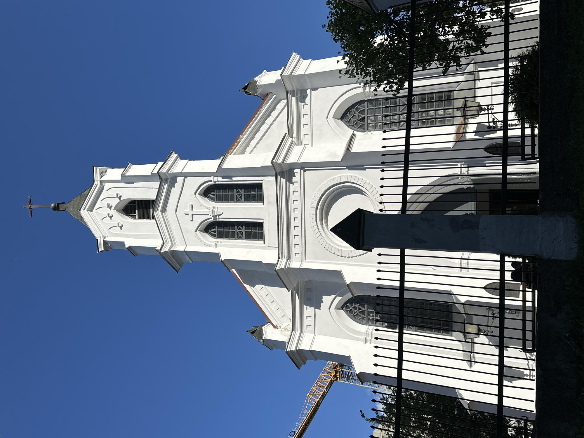 The image shows the facade of a white church with a tall bell tower, against a clear blue sky. The church has gothic-style arched windows and ornate architectural details. A black metal fence is in the foreground, partially obscuring the lower part of the building. To the left, a yellow construction crane is visible, suggesting ongoing development or renovation in the area. Green foliage from trees is seen to the right. The architectural style and the presence of a crane suggest this might be a historical building undergoing preservation or modernization, common in urban settings like Minsk, Belarus. The specific church is likely the Holy Spirit Cathedral, a prominent landmark in Minsk, known for its white facade and distinctive tower.