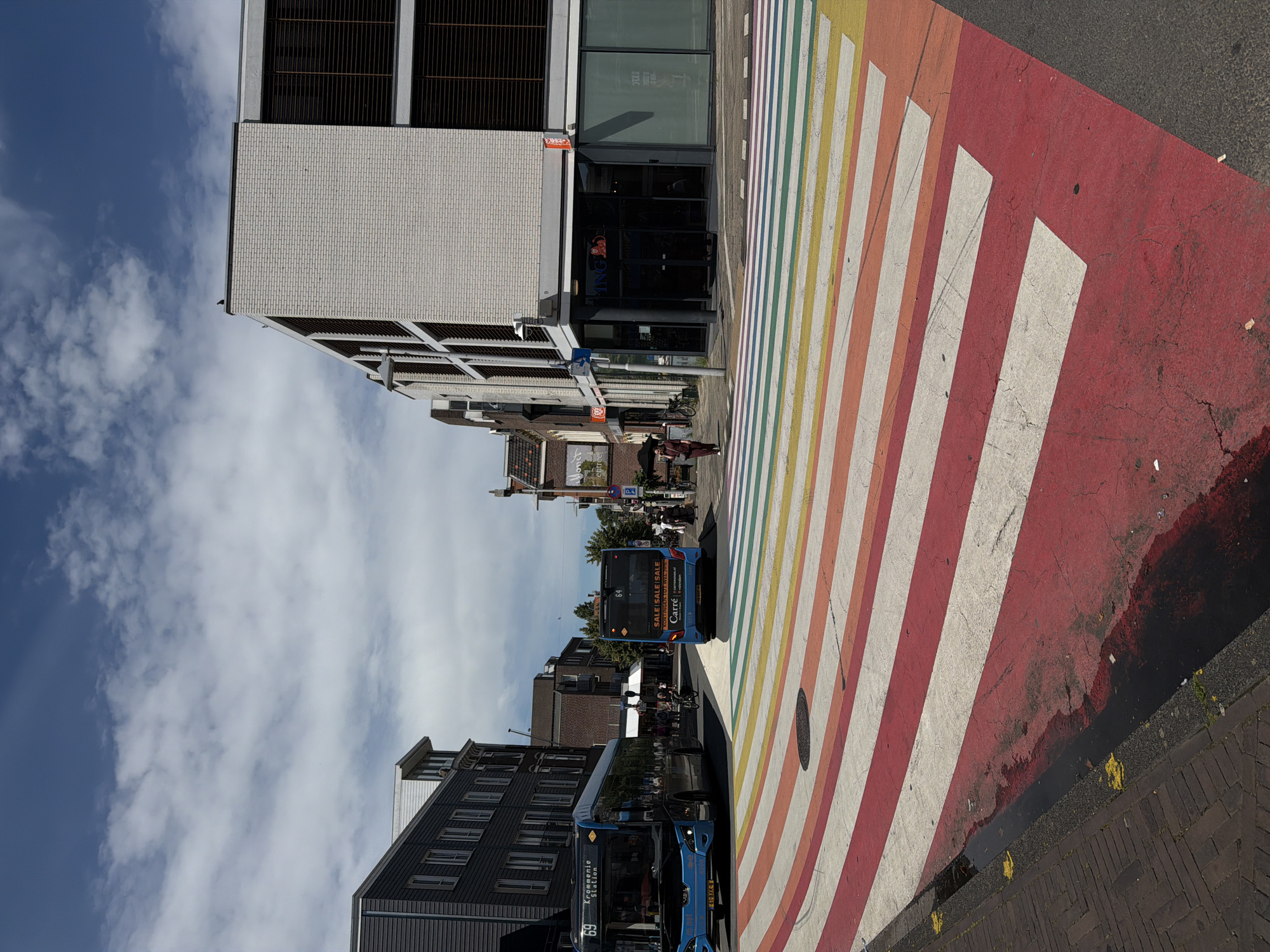 The image depicts a vibrant, rainbow-colored crosswalk in Zaandam, Netherlands. Two blue buses are present, one on the left and one on the right, with visible route information. The bus on the left shows "69 Krommenie Station" and the bus on the right shows "64 SALE SALE SALE CARRE". Several people are visible in the background, some walking and others cycling. The environment includes modern buildings constructed with brick and large windows, as well as older buildings with darker facades. The sky is blue with scattered clouds, indicating a daytime scene. The crosswalk itself is the dominant feature, painted with wide, colorful stripes of red, orange, yellow, green, blue, and purple. The scene appears to be a busy urban intersection, likely in the city center of Zaandam.