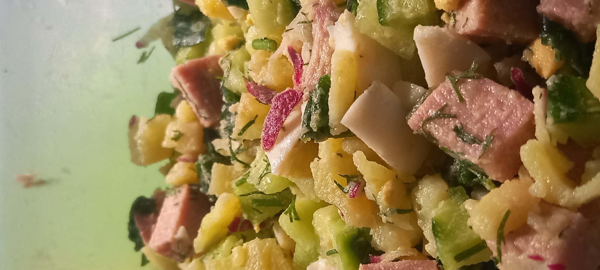The image shows a close-up view of a potato salad. The salad contains diced potatoes, cucumbers, hard-boiled eggs, and what appears to be diced ham or some other processed meat. Fresh dill and possibly radishes are also visible, adding color and texture. The salad is mixed with a creamy dressing. The background is a blurred, light green, suggesting it might be a plastic container or bowl. There are no discernible landmarks or elements that would help to locate this scene within the city of Kryvyi Rih, Ukraine. The image appears to be taken indoors, possibly in a kitchen or dining area. There are no people or animals present in the image. The focus is entirely on the food itself.