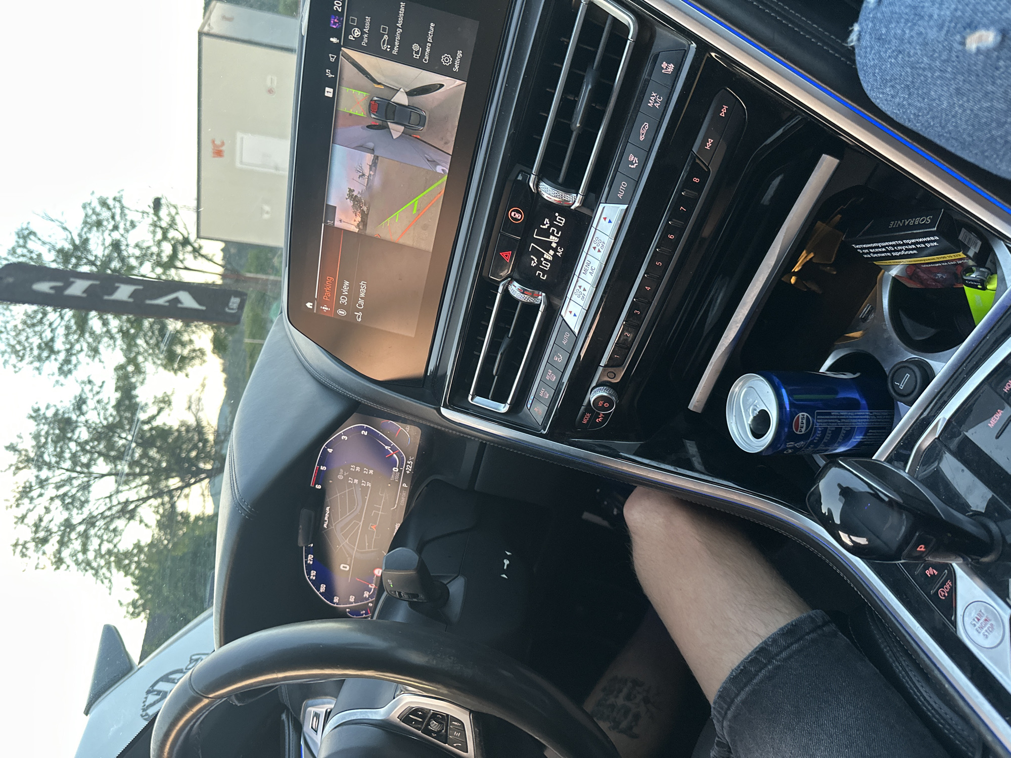 The image shows the interior of a car, likely a modern BMW based on the dashboard and infotainment system. A person's arm and leg are visible, suggesting they are in the driver's or passenger's seat. The car's digital dashboard displays navigation and vehicle information, and the central infotainment screen shows a parking camera view with graphics indicating distance and direction. A can of Pepsi is in the center console, next to a pack of cigarettes and a lighter. The exterior view through the windshield shows trees and a portable toilet structure labeled "WC," indicating an outdoor setting. A "VIP" banner is also visible outside the car. The time of day appears to be late afternoon or early evening, with warm light. There is no explicit information in the image to pinpoint the location in the city of Kostenets, Bulgaria, although the presence of a portable toilet and outdoor setting might suggest a public event or a rest stop.