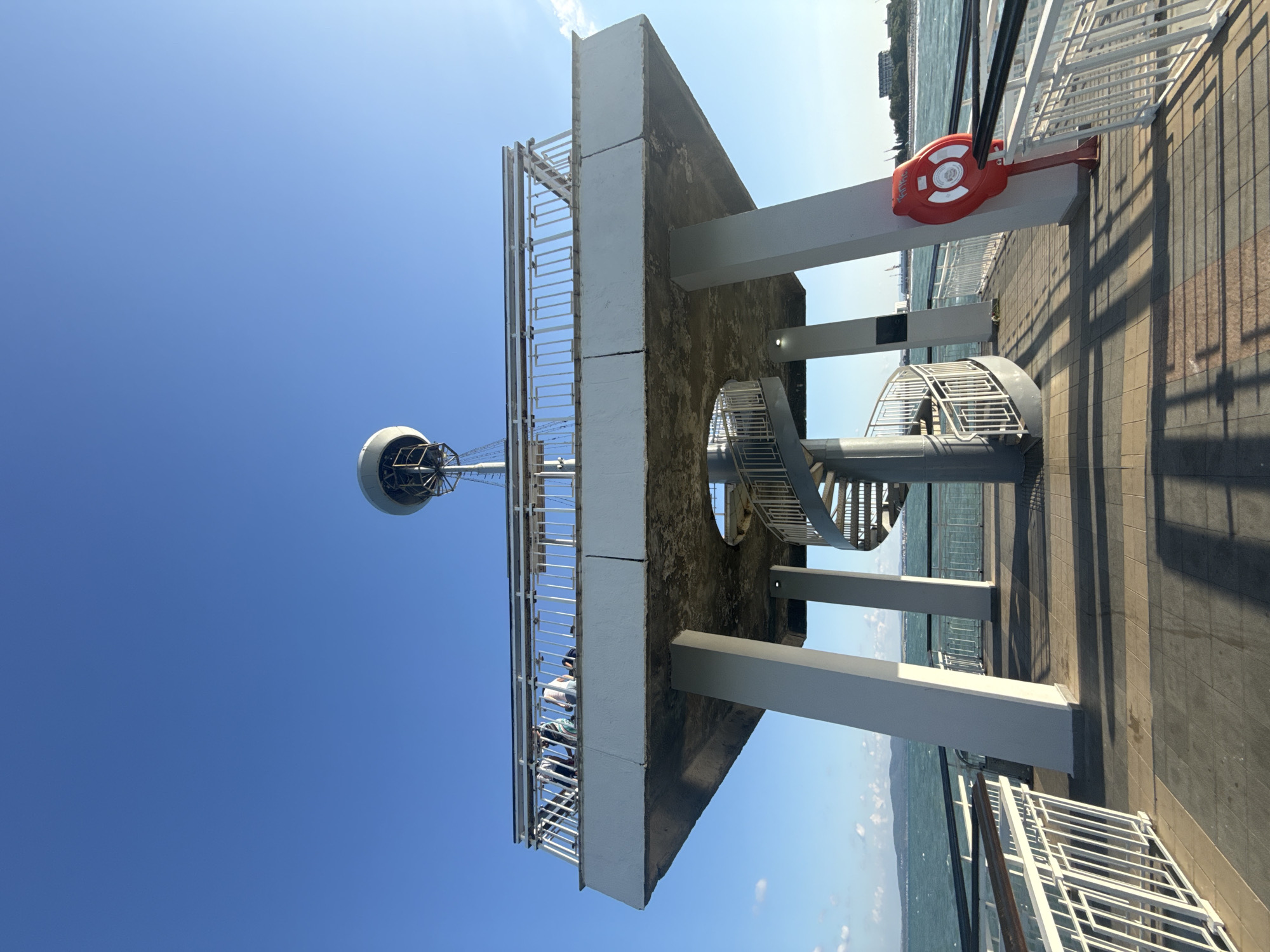 The image shows a pier with a spiral staircase leading up to a viewing platform. On the platform, several people are standing and looking out at the sea. A large, dome-shaped structure sits atop the platform, possibly a weather station or observation equipment. To the right, a red life preserver is mounted on a pillar. The pier itself is paved with tiles, and railings line the sides, providing safety. In the background, the blue sea stretches to the horizon, meeting a clear, bright sky. The sea is calm, with a few distant boats and buildings visible along the coastline. The pier is part of the Burgas Sea Garden, a prominent recreational area in Burgas, Bulgaria.