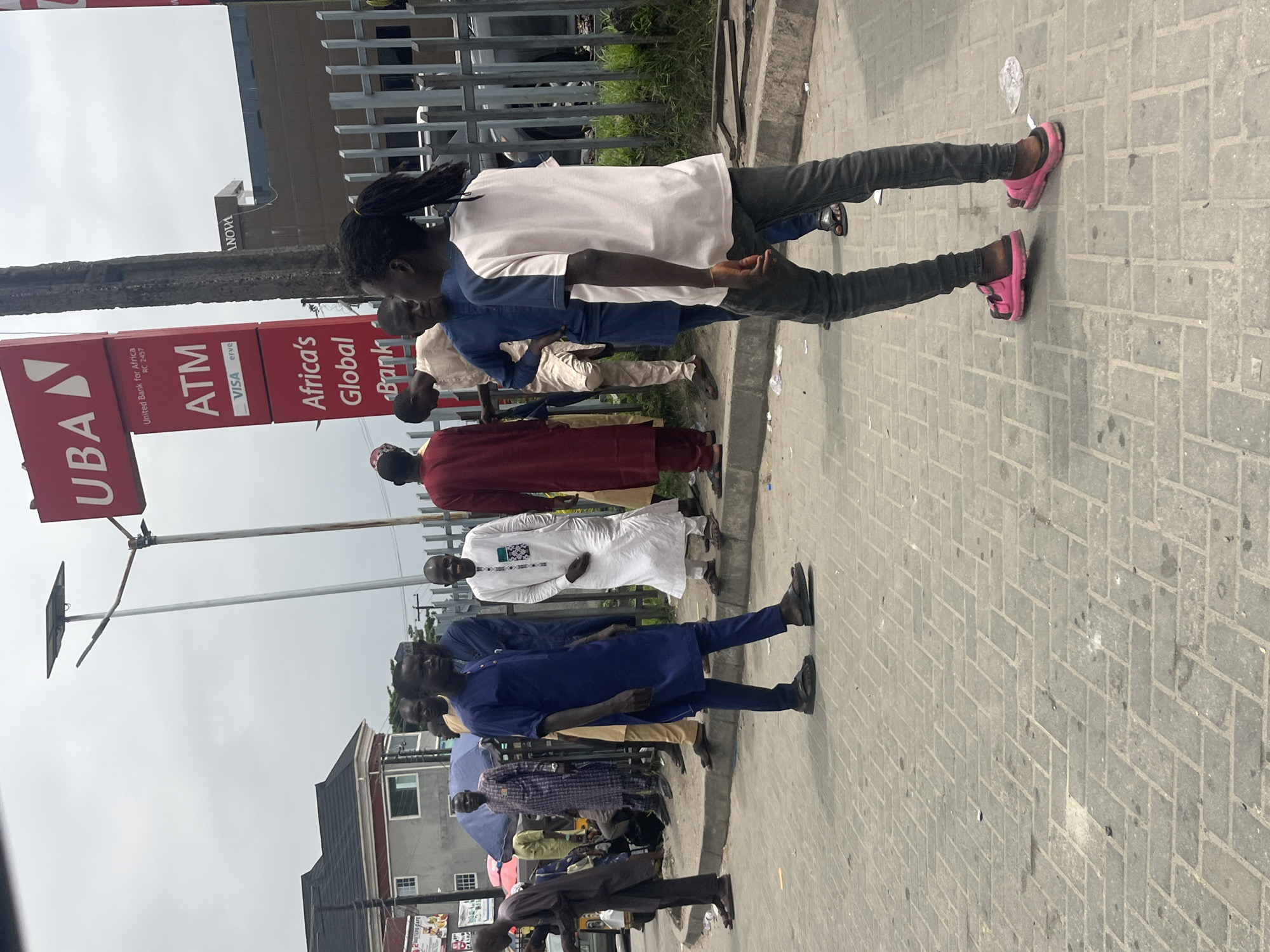 A medium shot captures a street scene with several individuals in what appears to be Ado, Nigeria. In the foreground, a young person with dark braided hair walks away from the camera, wearing a light-colored t-shirt and dark pants, with pink sandals. To their left, a man in a dark blue traditional outfit and dark sandals stands facing the viewer. Behind him, other men are visible in various traditional garments, including a man in a white kaftan. A red sign with white lettering for UBA (United Bank for Africa) and ATM is prominently displayed on a pole. The ground is paved with rectangular bricks. In the background, buildings, a metal fence, and greenery can be seen under an overcast sky.