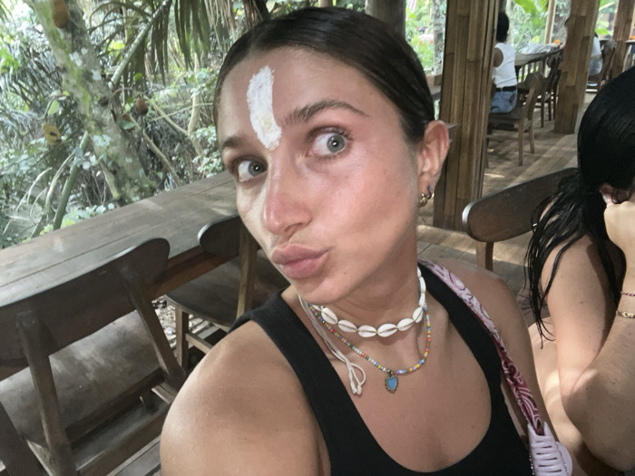 A woman is depicted in a close-up selfie taken in a semi-open-air establishment in Punggul, Indonesia. She has fair skin, dark hair pulled back, and a vertical white stripe of substance applied to her forehead. Her eyes are wide and directed at the camera, while her lips are pursed. She wears a black tank top, a cowrie shell necklace, and a multicolored beaded necklace with a blue-green pendant. Gold hoop earrings are visible on her right ear. A light-colored patterned strap is visible over her right shoulder. To her right, a second individual with dark hair is partially visible. In the background, wooden tables and chairs are arranged within a structure featuring bamboo or light wood pillars. Further back, another person in a white top is seated at a table. Lush green tropical foliage and trees are visible beyond the establishment, with several fruit-like objects hanging from branches on the far left.