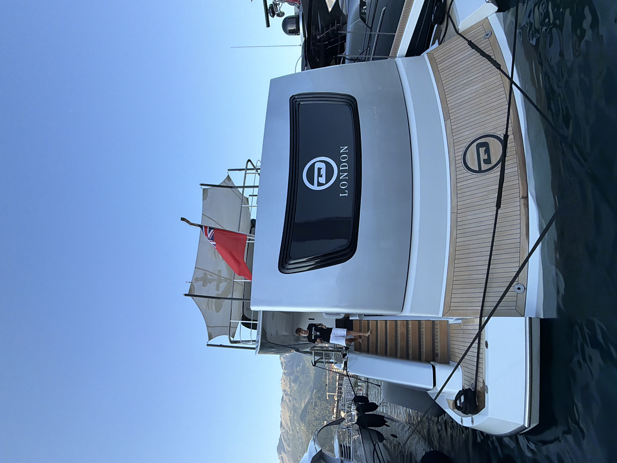 A young male wearing a black t-shirt with a logo and white shorts walks down wooden stairs on the side of a large, modern, silver yacht named "LONDON". The yacht is docked in a harbor in Budva, Montenegro, with mountains visible in the background under a clear blue sky. The yacht has a dark rectangular window with a white logo and the word "LONDON" printed below it. Another smaller yacht is docked to the left of the "LONDON" yacht. The water is dark and calm. The overall scene depicts a sunny day in a coastal city.