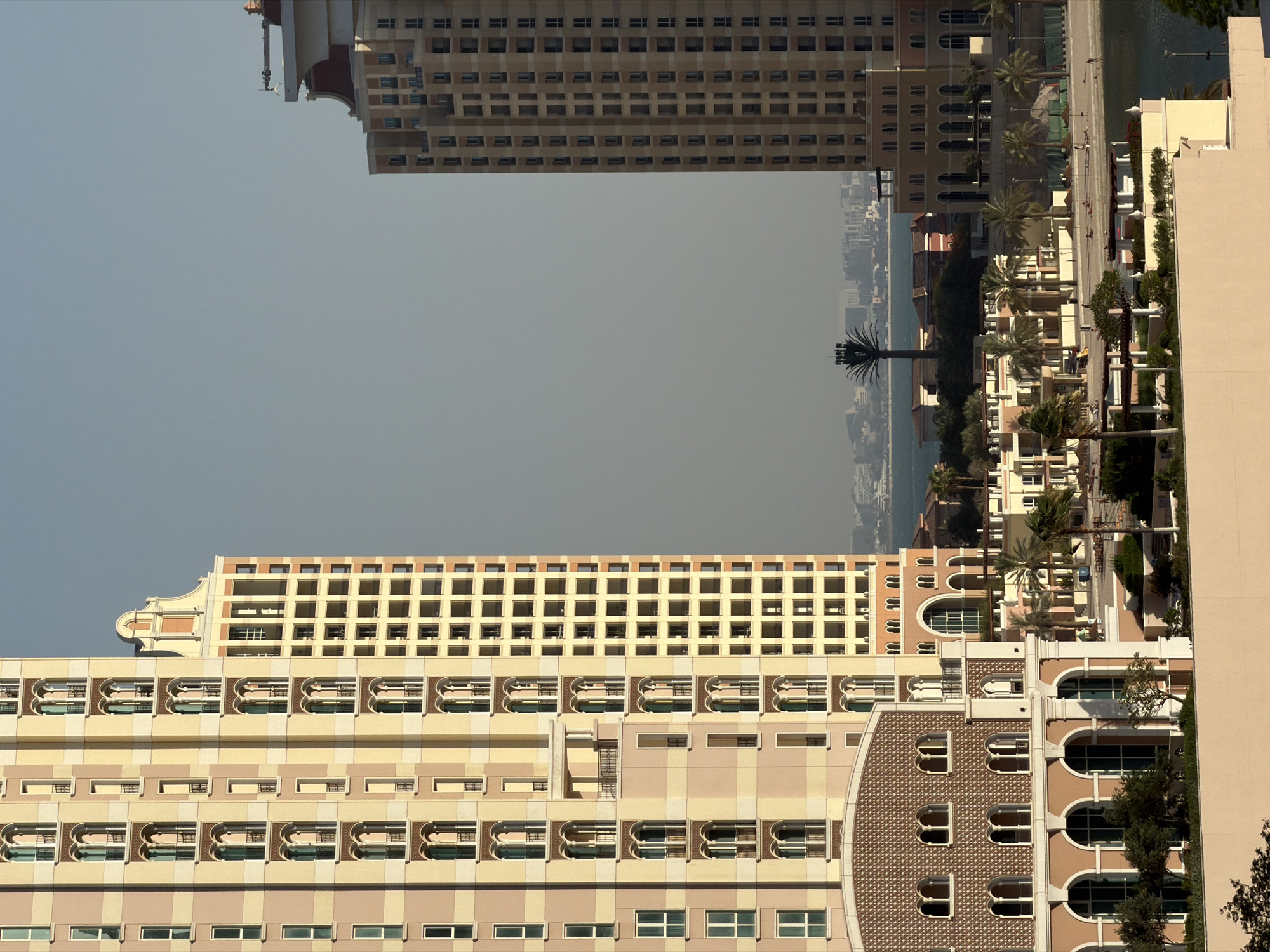 This image captures a modern, urban coastal scene dominated by two large, multi-story buildings and a body of water under a hazy sky. The photograph appears to have been taken in daylight, possibly late morning or early afternoon, with soft, diffused lighting.
In the foreground and spanning the left side, a large, ornate building rises, featuring multiple architectural styles across its facade. Its lower sections are composed of light beige and brown blocks with rectangular windows. Higher up, there are several rows of windows with distinctive arched or scalloped frames, often arranged in groups. A prominent section of the building exhibits a curved, reddish-brown tiled facade with multiple arched windows.
On the right side, a second, taller building stands with a more uniform, grid-like pattern of numerous rectangular windows. It features decorative elements on its roofline. Between these two prominent structures and extending towards the background is a waterfront area. This area is lined with a pathway or promenade, dotted with numerous green palm trees and smaller, light-colored buildings with balconies and arched doorways.
Beyond the waterfront and trees, a wide expanse of calm, blue-grey water stretches towards the horizon. In the far distance, a faint outline of a hazy cityscape or landmass is visible. The sky is a uniform, light grey-blue, suggesting either overcast conditions or a hazy atmosphere, contributing to the soft light. There are no people or specific activities discernible in the image. The architectural style, particularly the arches and light-colored facades, along with the palm trees, suggest a location in a warm climate, possibly the Middle East or a similar coastal region known for modern development. No visible text is present.