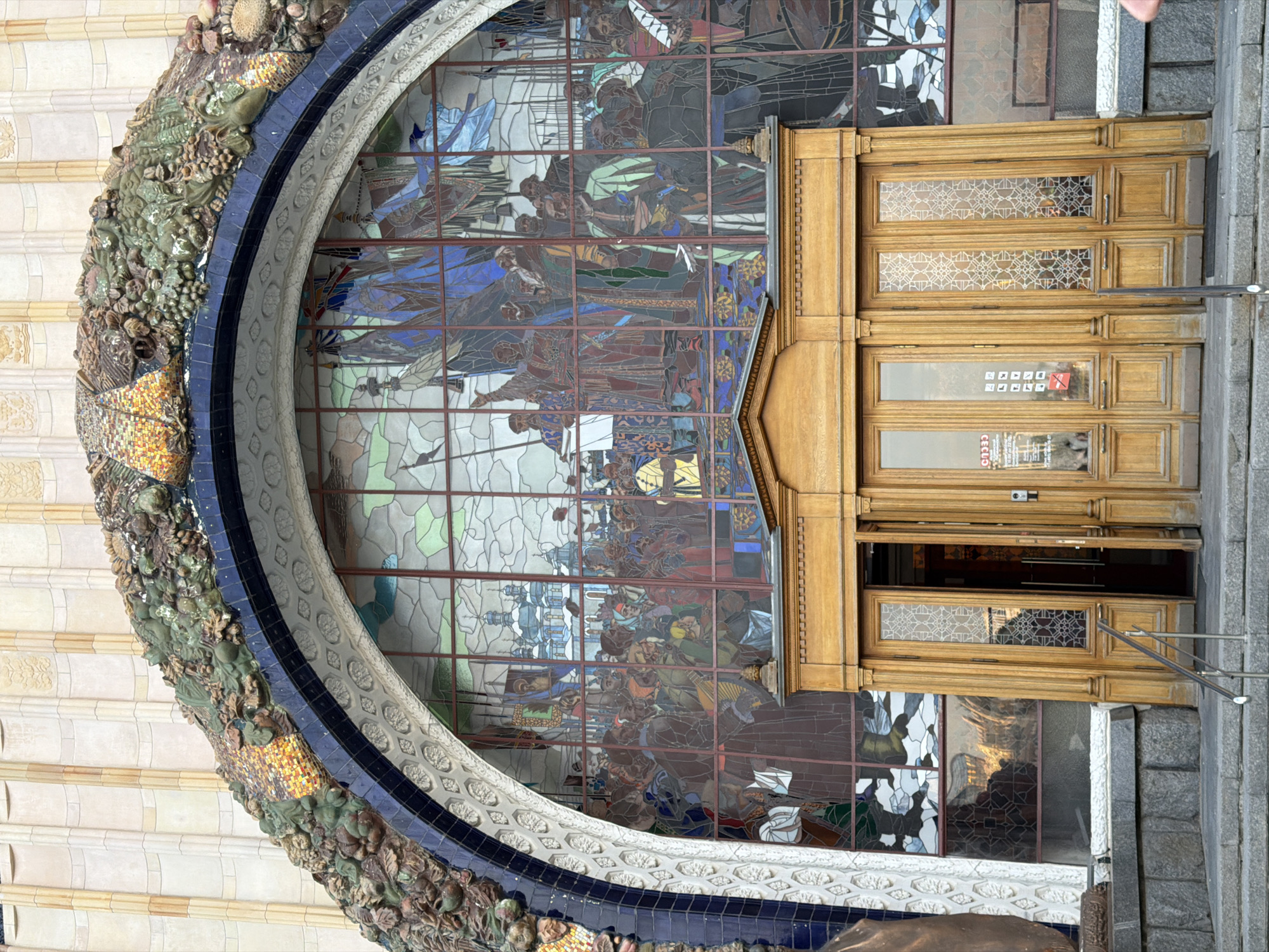 The image shows the facade of a building in Moscow, Russia, specifically an entrance to the VDNKh metro station. The entrance features a large arched window with a stained-glass mural depicting historical or allegorical scenes with figures in various colors. The arch is framed by ornate decorations including sculpted vegetation and a blue-tiled border. To the right of the arch is a set of wooden doors, some of which are open, revealing a glimpse inside the station. The doors are decorated with geometric patterns. The building is constructed from light-colored stone and appears to be well-maintained.