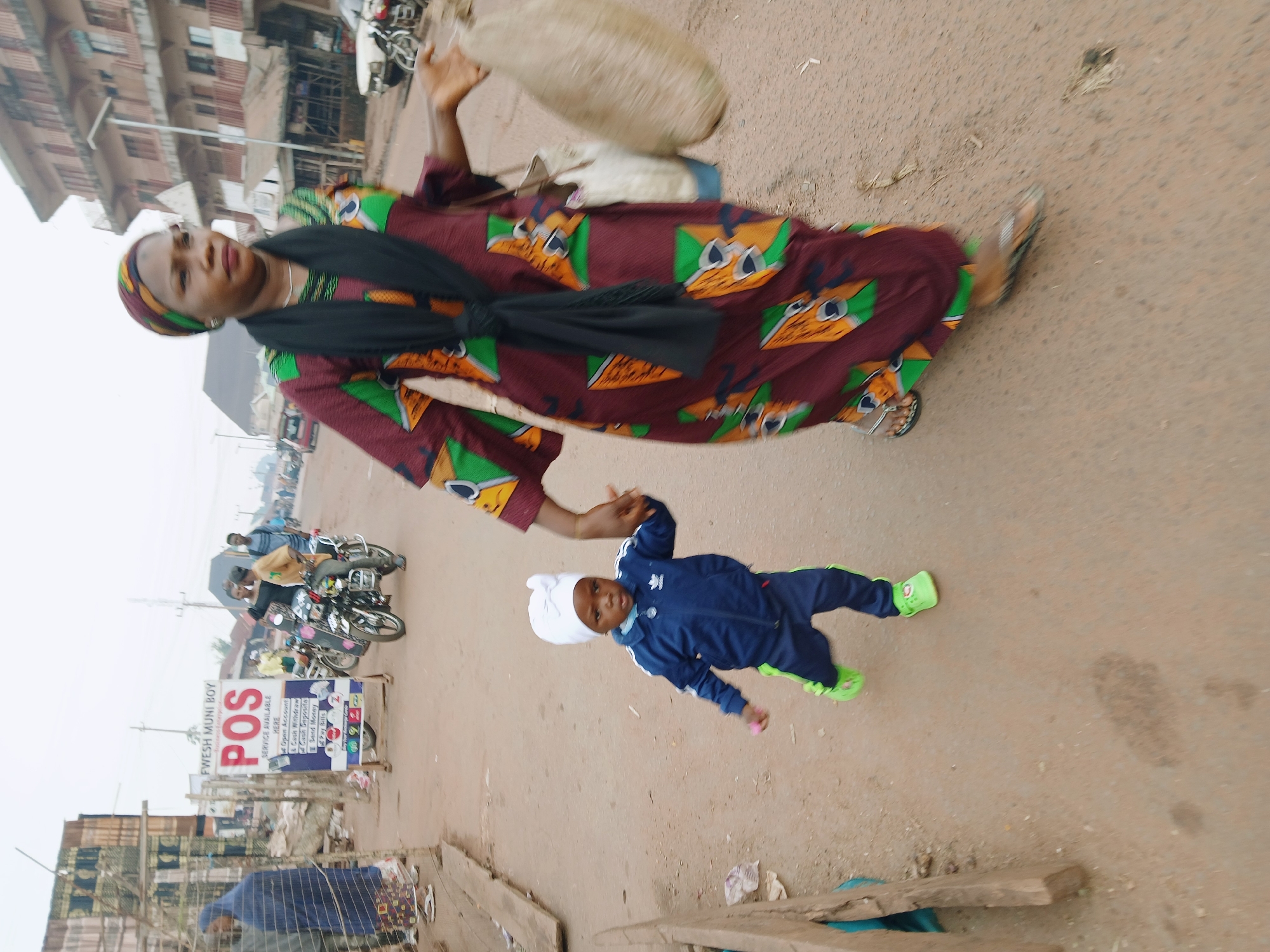 In Ankpa, Nigeria, a medium shot reveals a woman and a young child on a street, with the image rotated for an unusual perspective. The woman, wearing a patterned maroon dress with green and orange giraffe-like figures, and a black sash, carries a beige sack atop her head and holds the hand of a toddler dressed in a blue tracksuit and light green shoes.
In the background, a "POS SERVICE AVAILABLE HERE" sign is visible, indicating a mobile money service. Motorcycles and buildings are also present, suggesting a busy urban environment. The street is a sandy, off-white color. The overall environment appears somewhat desolate, with visible trash and wooden planks scattered in the foreground.
