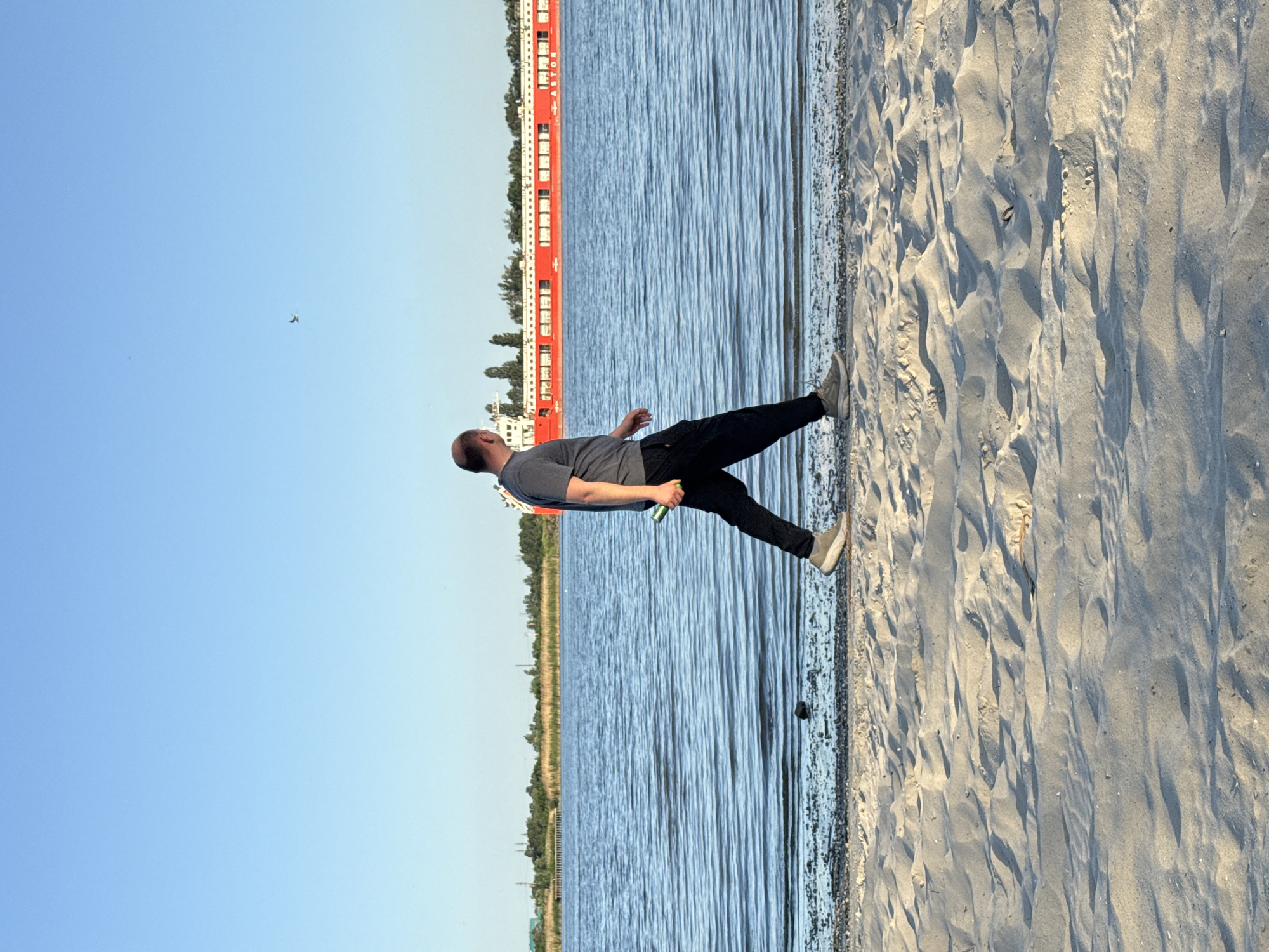 A man stands on a sandy beach along a river in Rostov-on-Don, Russia. The beach is to the right, with the river extending to the left. The man is wearing a gray shirt and black pants, and is holding a beverage can in his right hand. A large red barge is visible on the river. The sky is clear and blue.