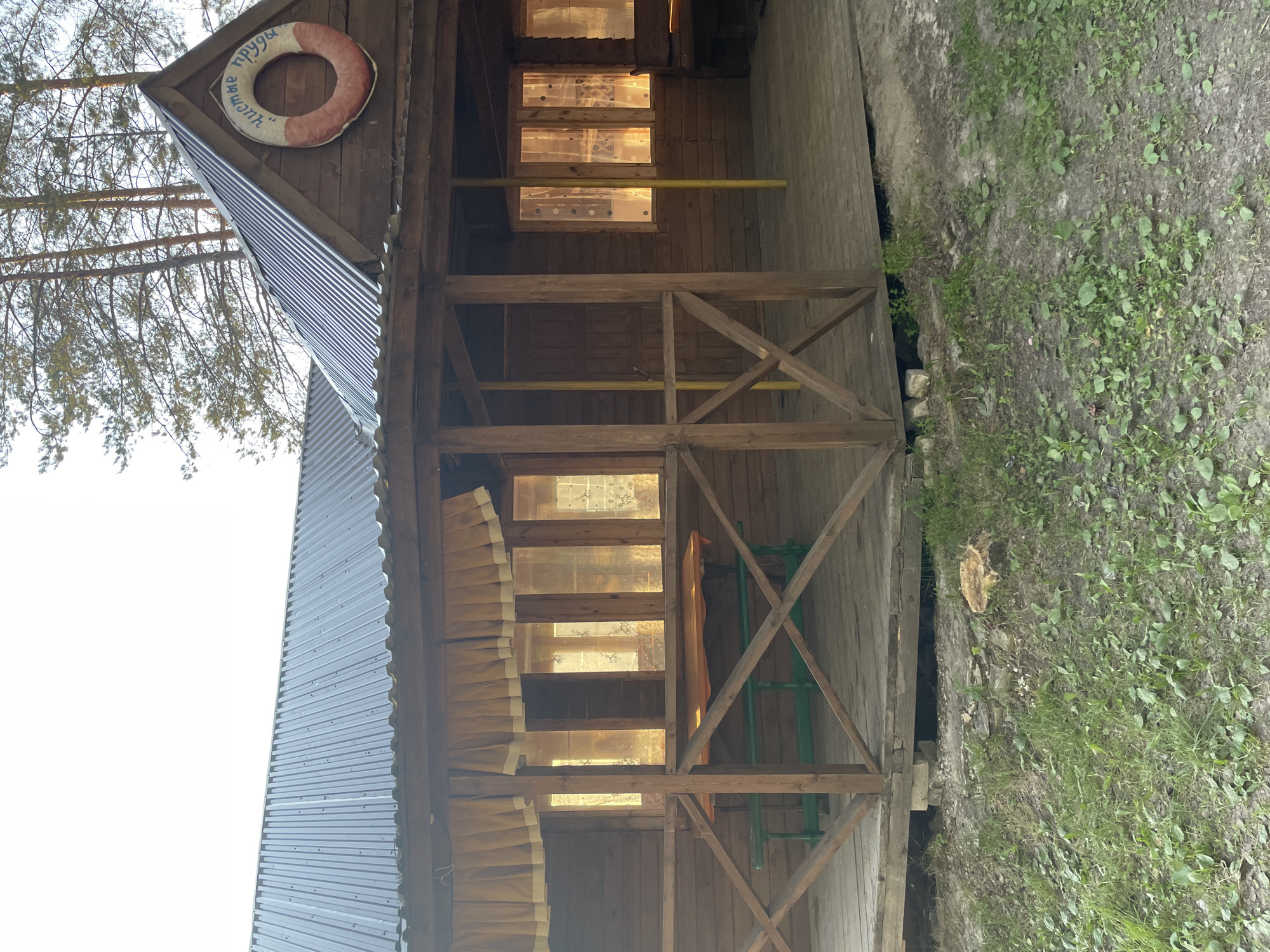 The image depicts a wooden cabin with a corrugated metal roof, situated in a rural or natural setting, likely near a body of water, given the sign on the lifebuoy reading "Чистые пруды" (Chistye Prudyi), which translates to "Clean Ponds". The cabin has a covered porch with wooden railings and a wooden deck. Through the windows, which are covered by yellow curtains, the interior appears to be illuminated. There is outdoor furniture, including a picnic table with benches, visible on the porch. The surrounding environment is characterized by grass and trees, suggesting a park or recreational area. The scene is captured in daylight, with sunlight casting a warm glow through the windows.