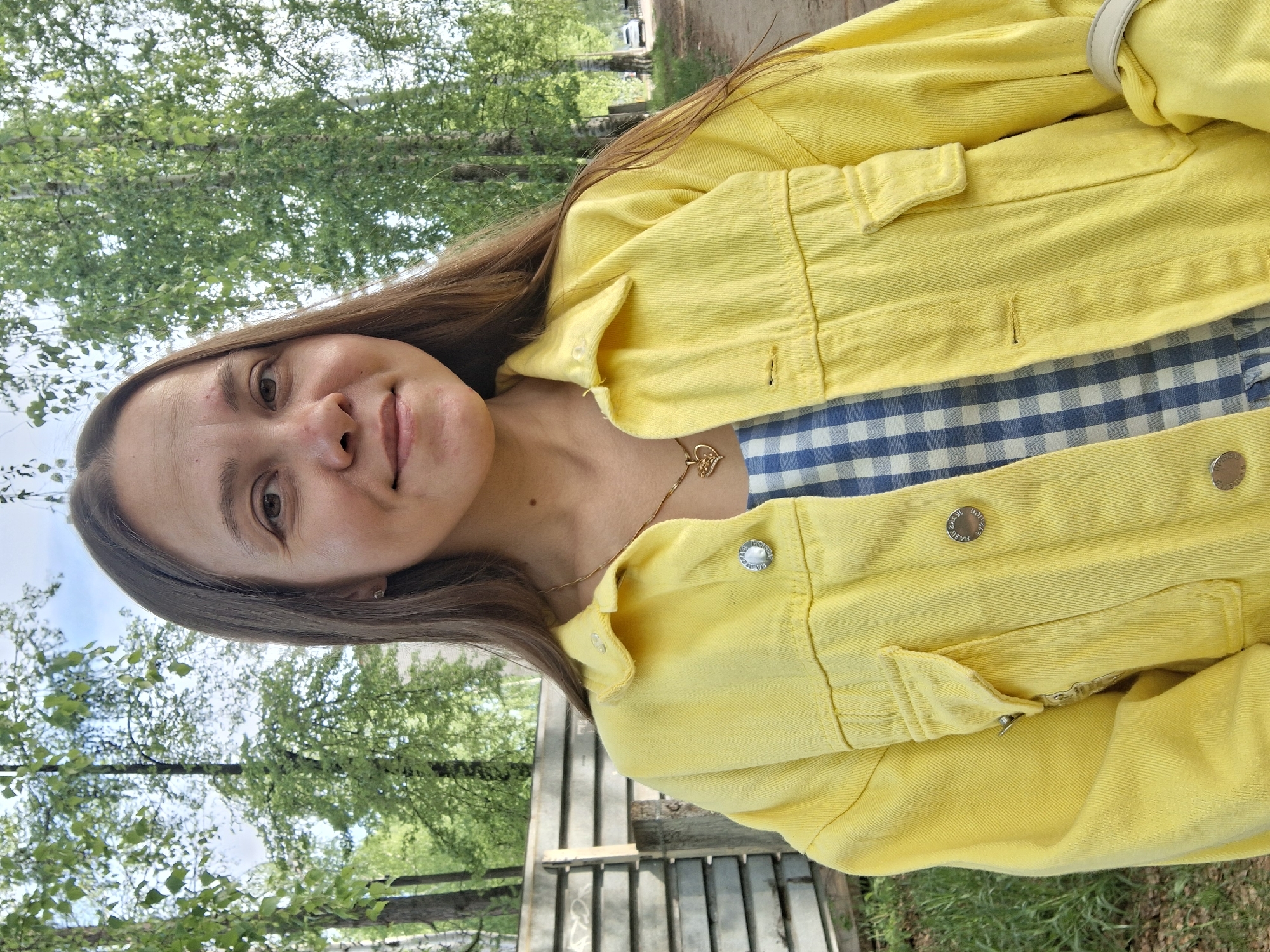 A woman with long brown hair, wearing a yellow denim jacket over a blue and white checkered top, is photographed outdoors. She is smiling slightly at the camera. The background includes trees and a wooden fence, suggesting a park or natural area. A gold necklace with a pendant is visible. Based on the geotag information, the location is in Chaikovsky, Russia.