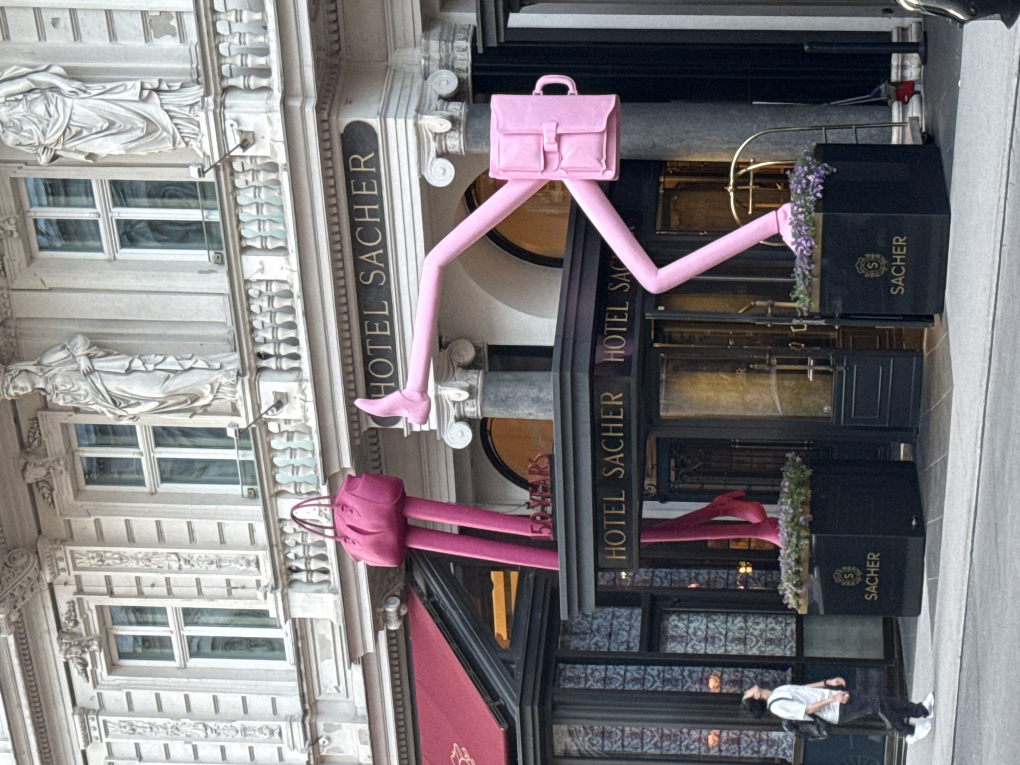 This image captures a street-level view of the historic Hotel Sacher in Vienna, Austria, adorned with whimsical art installations. The primary subject is the hotel's ornate, classical facade, characterized by light-colored stone, multiple large windows, decorative balconies, and intricate carvings, including draped classical statues.
Dominating the central part of the image are two large, stylized sculptures of bags with legs. The upper sculpture is a bright pink briefcase or satchel, with long, slender legs and small feet, appearing to stride dynamically over the main entrance of the hotel. Below it, over a red awning, is a second, darker pink sculpture resembling a handbag, also equipped with similar long legs.
The hotel entrance features large dark-framed windows and doors, flanked by black planter boxes containing purple flowers. Each planter box clearly displays the "SACHER" name with an "S" logo. A prominent sign vertically mounted on the building reads "HOTEL SACHER," with other instances of the hotel's name visible on signage and below the upper sculpture, where "150 YEARS" can also be partially seen.
In the bottom right, a person dressed in a white t-shirt, dark pants, and white shoes stands on the sidewalk, looking up at the hotel and the sculptures, suggesting engagement with the art. The lighting indicates it is daytime. The overall scene presents a blend of historical architecture and modern, playful art.