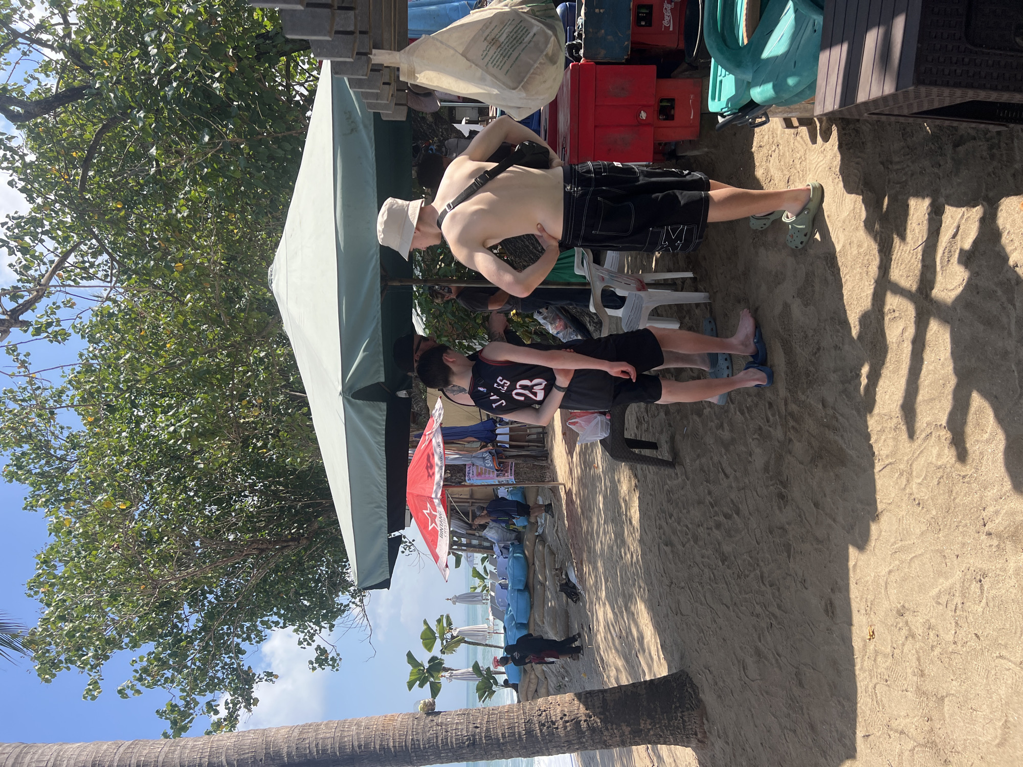 A group of people are gathered on a sandy beach in Kuta, Indonesia. A tall palm tree is on the left side of the frame, casting a shadow on the sand. In the background, the ocean is visible with waves crashing. Two young men are standing near a stall with a large green umbrella. One is wearing a black tank top with the number 23 and shorts, and the other is wearing shorts and a bucket hat. Another person in a black shirt and shorts is standing behind them. There are several blue beanbag chairs set up on the sand, and a red umbrella with the Bintang logo is also visible. A vendor stall with various items is set up under the green umbrella, with red crates and coolers nearby. Further down the beach, several people are walking towards the water. The sky is clear and blue with some clouds.
