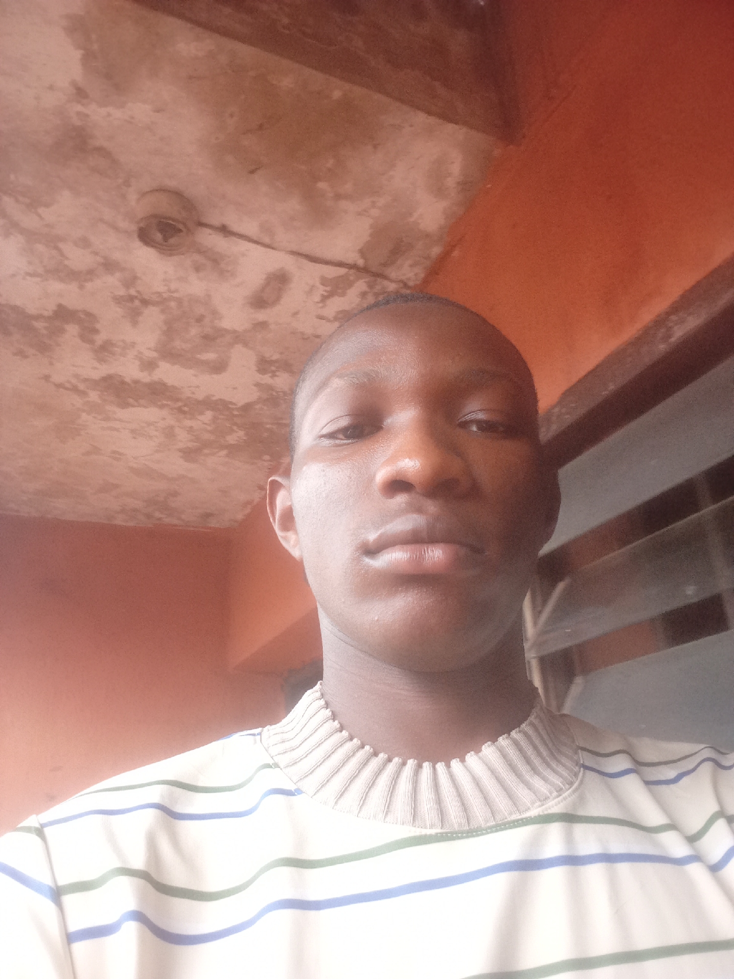 The image is a close-up, waist-up, selfie-style photograph taken indoors, likely in Onitsha, Nigeria. The subject is a young Black male facing the camera. He has dark skin, short-cropped hair, and is wearing a cream-colored, long-sleeved shirt with horizontal stripes of light green and blue. The shirt has a ribbed, mock-neck collar.
The background is indistinct but appears to be an interior space with an orange-toned wall and a ceiling that shows signs of wear and a light fixture. To the right of the subject, a window with horizontal blinds is partially visible. The lighting suggests an indoor setting, possibly with natural light from a window. The overall impression is casual and personal.