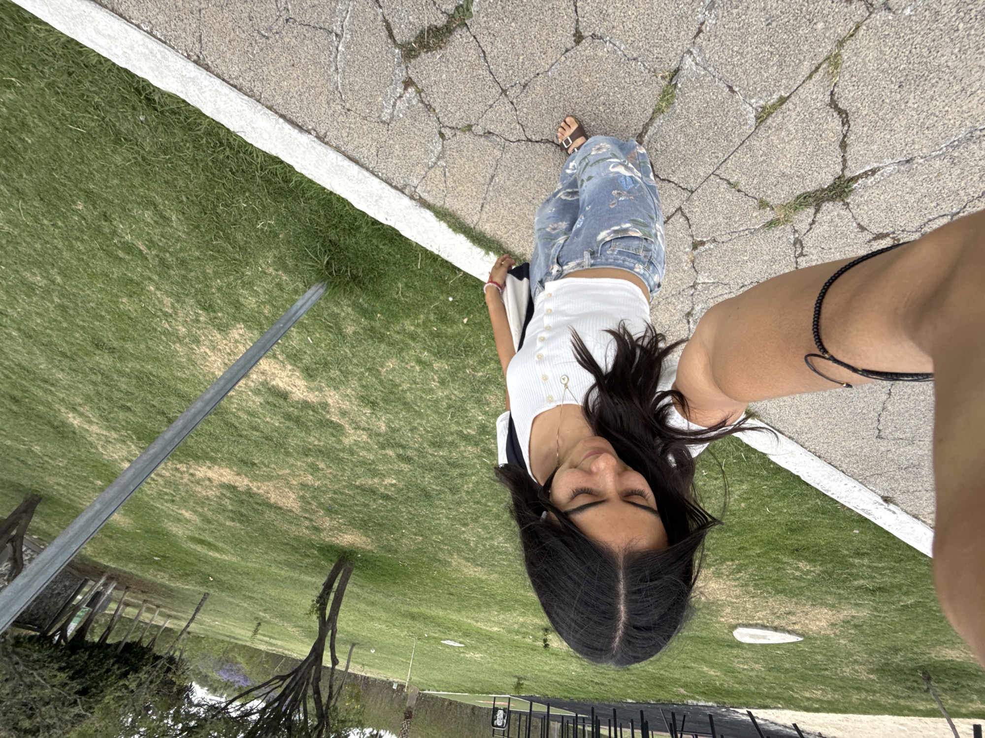 The image shows a young woman standing on a cracked concrete walkway next to a patch of grass. She is wearing blue patterned pants, a white top, and sandals. Her long, dark hair falls around her face. In the background, there is a grassy area with trees, a fence, and what appears to be a metal pole or structure. The angle suggests the photo was taken from a high vantage point or with a phone held at an unusual angle. The photo was taken outdoors during the daytime in Puebla, Mexico.