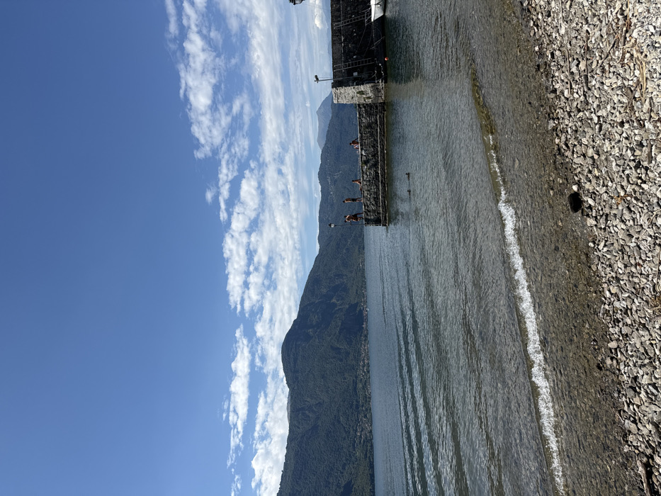 The image captures a sunny daytime scene on the shore of Lake Como in Abbadia Lariana, Italy. The foreground features a pebbled beach with a small wave breaking on the immediate shoreline. The lake water is calm with noticeable ripples, appearing clear near the shore and darker blue-green further out.
In the mid-ground, a dark stone pier or breakwater extends into the lake from the right side of the frame. It is constructed of large, dark grey stones and has a flat surface. Five individuals are present around this structure. On the pier, two individuals stand near the edge, one appearing to be preparing to enter the water. A third person is seated further back on the pier. Two additional individuals are visible in the water, partially submerged, swimming or floating near the pier.
To the right of the pier, a small white boat with a pointed bow is moored to the stone structure. A dark pole or light fixture is visible on the far right end of the pier.
The background is dominated by steep, forested mountains rising directly from the far side of the lake, extending across the entire horizon. A small village or settlement is discernible at the base of the mountains on the distant shore. The sky above is clear and bright blue with a band of scattered white cumulus clouds across the middle.