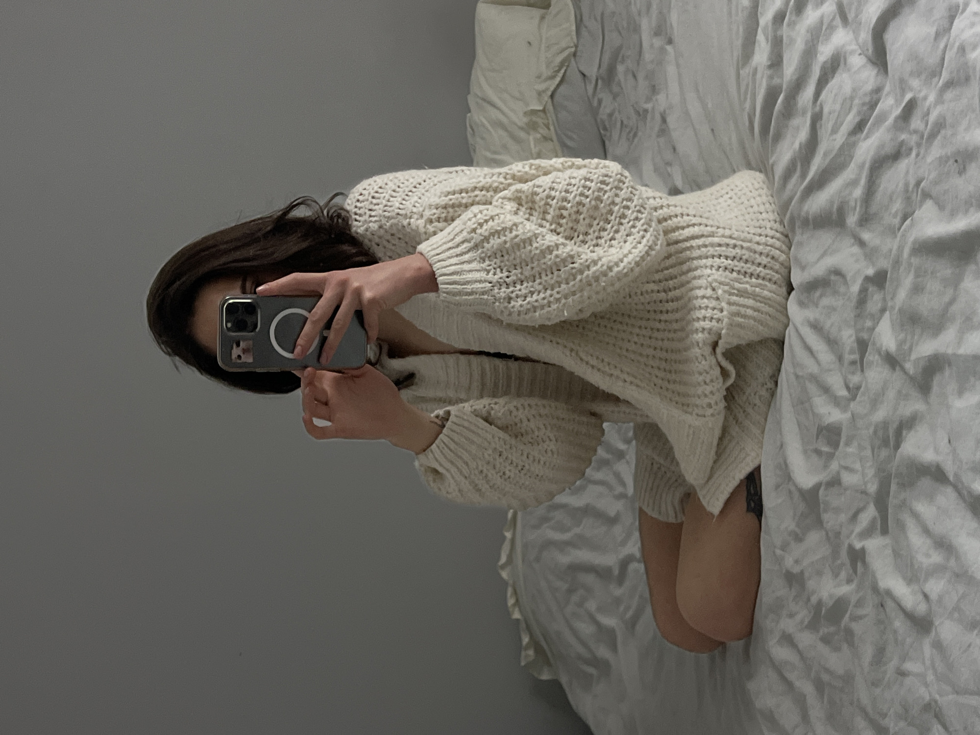 A person with dark hair sits cross-legged on a bed covered with white, wrinkled sheets. The person is wearing an oversized, cream-colored knit sweater. They are holding a smartphone up, taking a picture of themselves in a mirror, which is not visible in the frame. The background is a plain gray wall. There is no indication of the city of Ternopil, Ukraine.