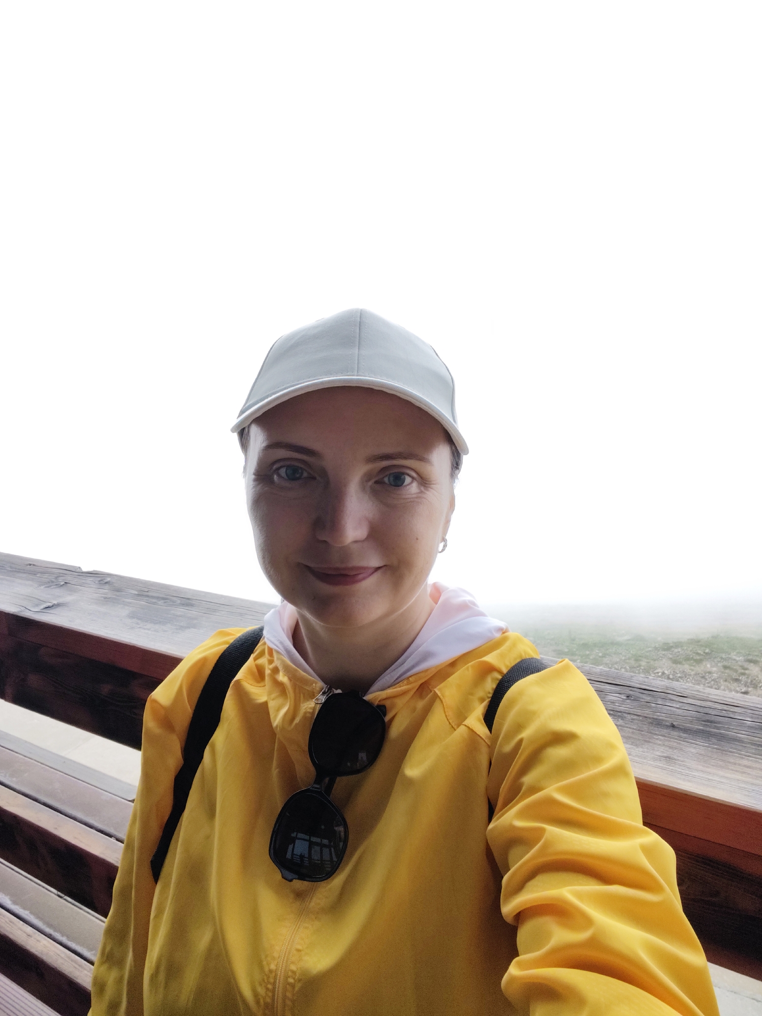 A woman wearing a light-colored baseball cap and a bright yellow hooded jacket is captured in a selfie. The jacket has a white trim on the hood and black sunglasses are clipped to the front zipper. She is sitting on a wooden bench with a wooden railing in front of her. The background is a hazy, foggy landscape with hints of green grass peeking through. The weather appears overcast and foggy, suggesting a cool or damp atmosphere. The woman has fair skin and blue eyes, and she is smiling gently at the camera. Her pose and the overall setting suggest a casual outing or travel experience, possibly in a mountainous or natural area given the fog. The image evokes a sense of calm and perhaps a bit of mystery due to the obscured background. The location is specified as Estosadok, Russia.