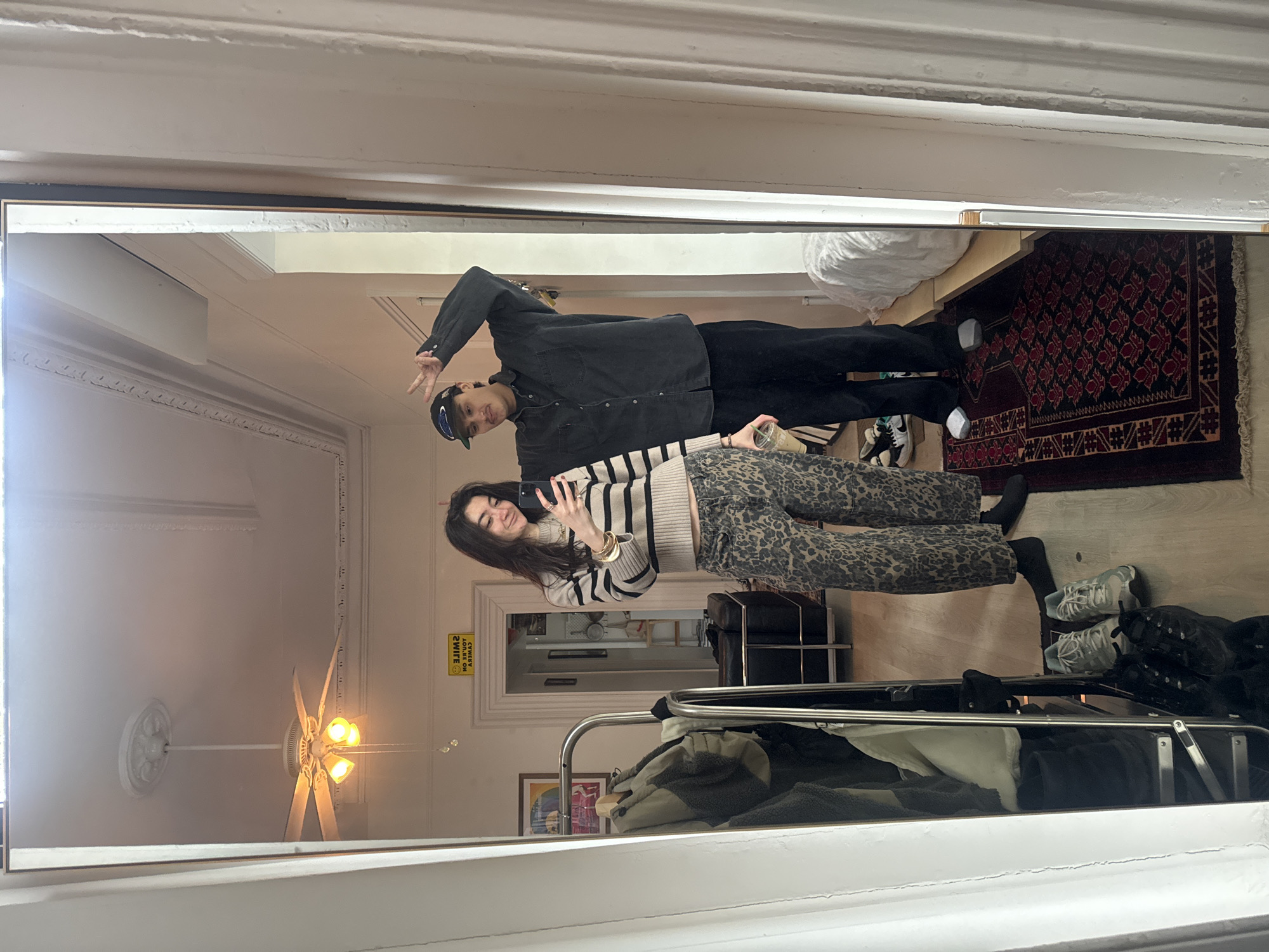 Two people are reflected in a mirror in what appears to be a room in an apartment. The woman is holding a smartphone, suggesting they are taking a selfie. She is wearing a striped sweater and patterned pants. The man beside her is wearing a dark jacket and baseball cap, and is gesturing with his hand. The room has a ceiling fan, decorative moldings, and a rug. A clothing rack is in the foreground, reflecting off the mirror.
Given the architectural details and the urban setting depicted in the reflection, it is plausible that the location is in New York City, United States of America.