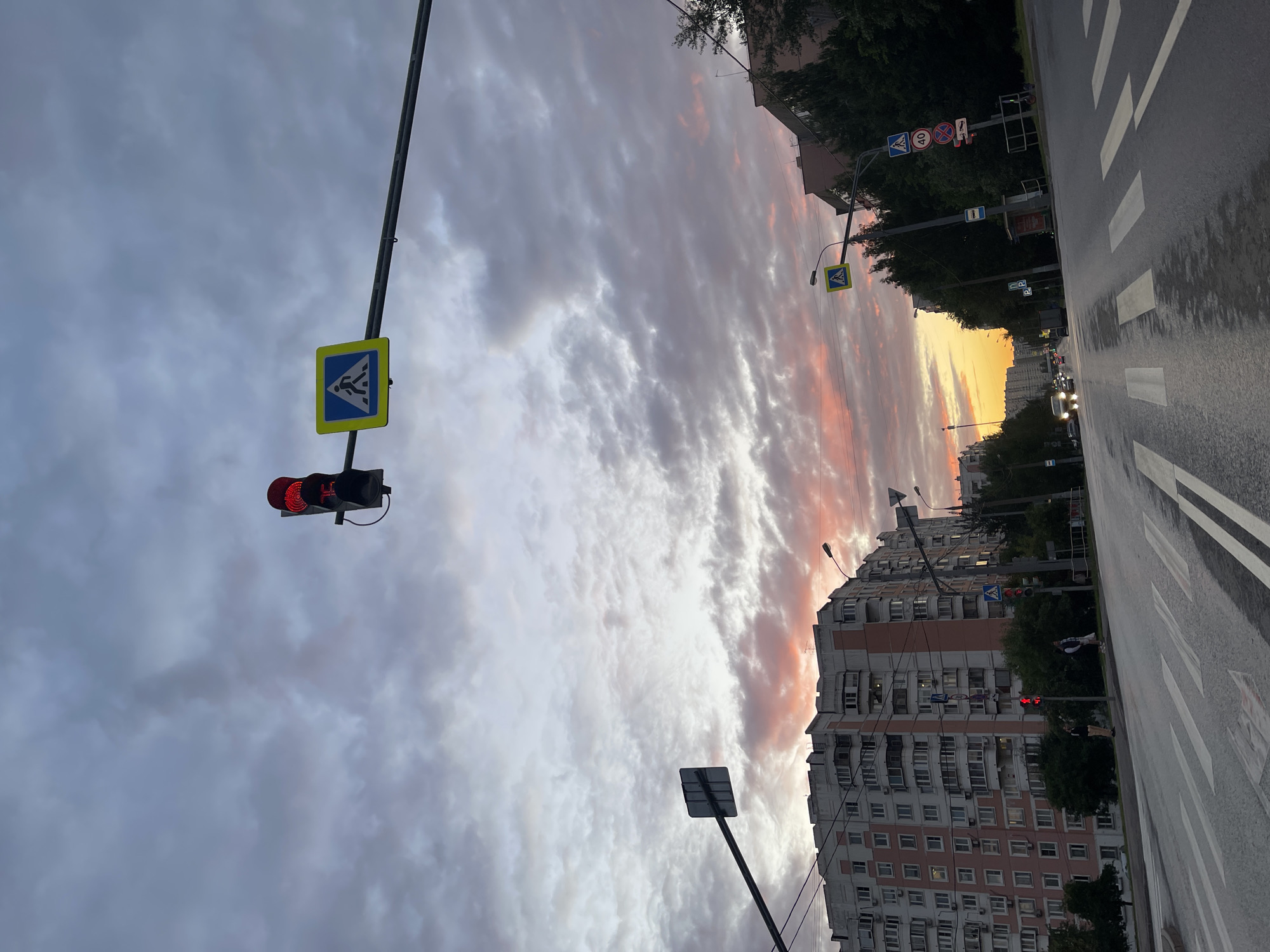 This image depicts a street scene in Moscow, Russia, during a cloudy sunset. The sky is filled with dramatic clouds streaked with hues of pink and orange from the setting sun. A tall, multi-story apartment building with many windows dominates the right side of the frame, characteristic of Soviet-era architecture. A wet asphalt road with a pedestrian crossing is visible in the foreground, indicating recent rain. Traffic lights and street signs are present, including a pedestrian crossing sign with a blue background and white figures, and a speed limit sign showing "40". The overall atmosphere is calm, with a touch of urban melancholy. Two individuals are visible crossing the street near a traffic light. The time of day is clearly evening, as indicated by the sunset colors and the red traffic light.