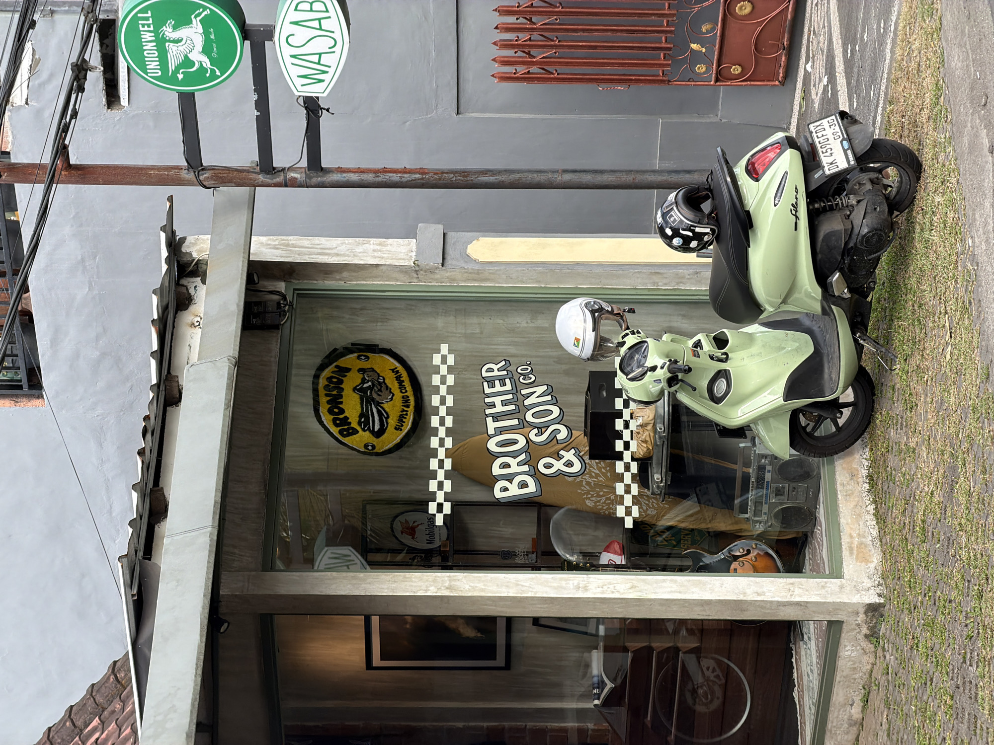 A light green scooter is parked on a sidewalk outside a shop. The shop's window has a "Bronson Supply and Company" logo and "Brother & Son Co." written on it. Signs for "Unionwell" and "Wasab" are above the shop. The scooter's license plate reads "DK 459GFDX." The location is North Kuta, Indonesia.