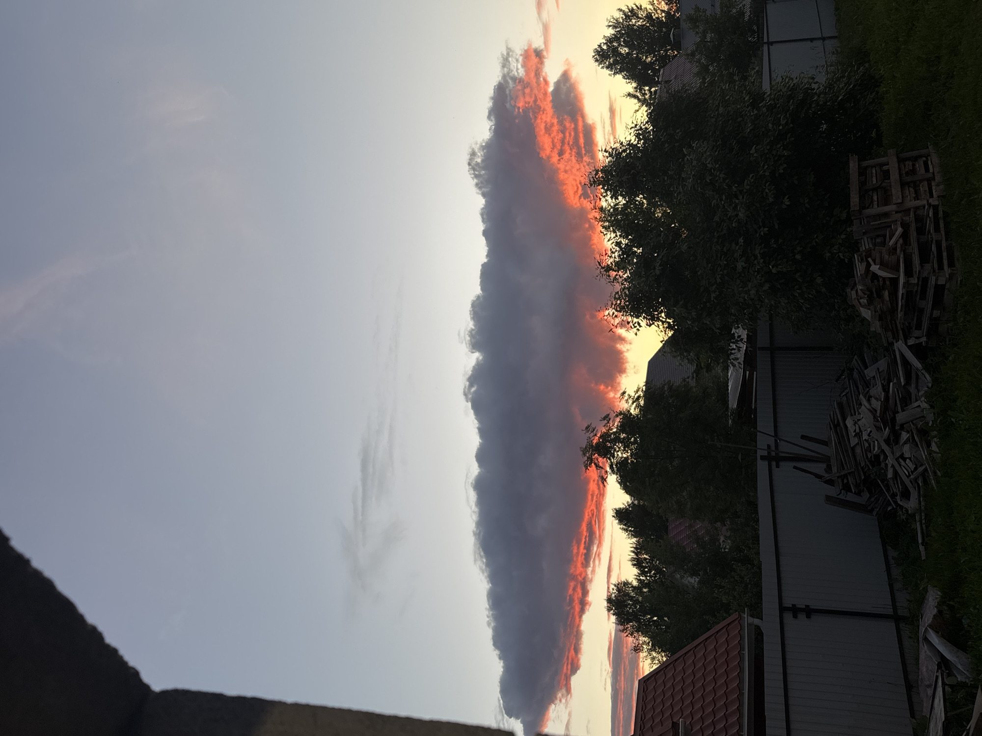 The image captures a sunset scene in Perm, Russia, with a dramatic cloud formation dominating the sky. A large, dark cloud is illuminated from below by fiery orange and red light, suggesting the sun is low on the horizon and casting its light through the cloud's undersides. The sky above transitions from a pale blue to a lighter hue near the horizon.
In the foreground, a dark, possibly concrete structure juts in from the left edge, framing the sky. Below the cloud, a line of trees with dense green foliage is silhouetted against the colorful sky. The tops of houses with red tiled roofs are visible peeking through the trees. A grey metal fence runs horizontally across the mid-ground, separating the residential area from a backyard. Piled wooden debris and pallets are scattered on the grass in the lower right portion of the image, indicating a storage or construction area. The overall impression is a blend of natural beauty and hints of human activity in a suburban or rural setting.