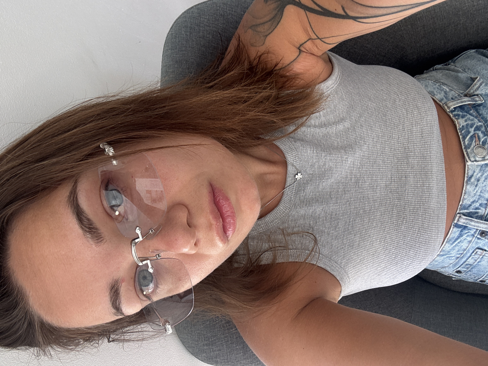 A close-up selfie of a woman with brown hair and blue eyes is shown. She is wearing a grey tank top, blue jeans, and clear rimless glasses. A small silver necklace with a clover charm is visible around her neck. She is sitting in a grey chair against a white wall, which is consistent with interior shots taken in apartments or hotels in Budva, Montenegro. Her left arm has a tattoo. The lighting suggests it is daytime, possibly indoors.
