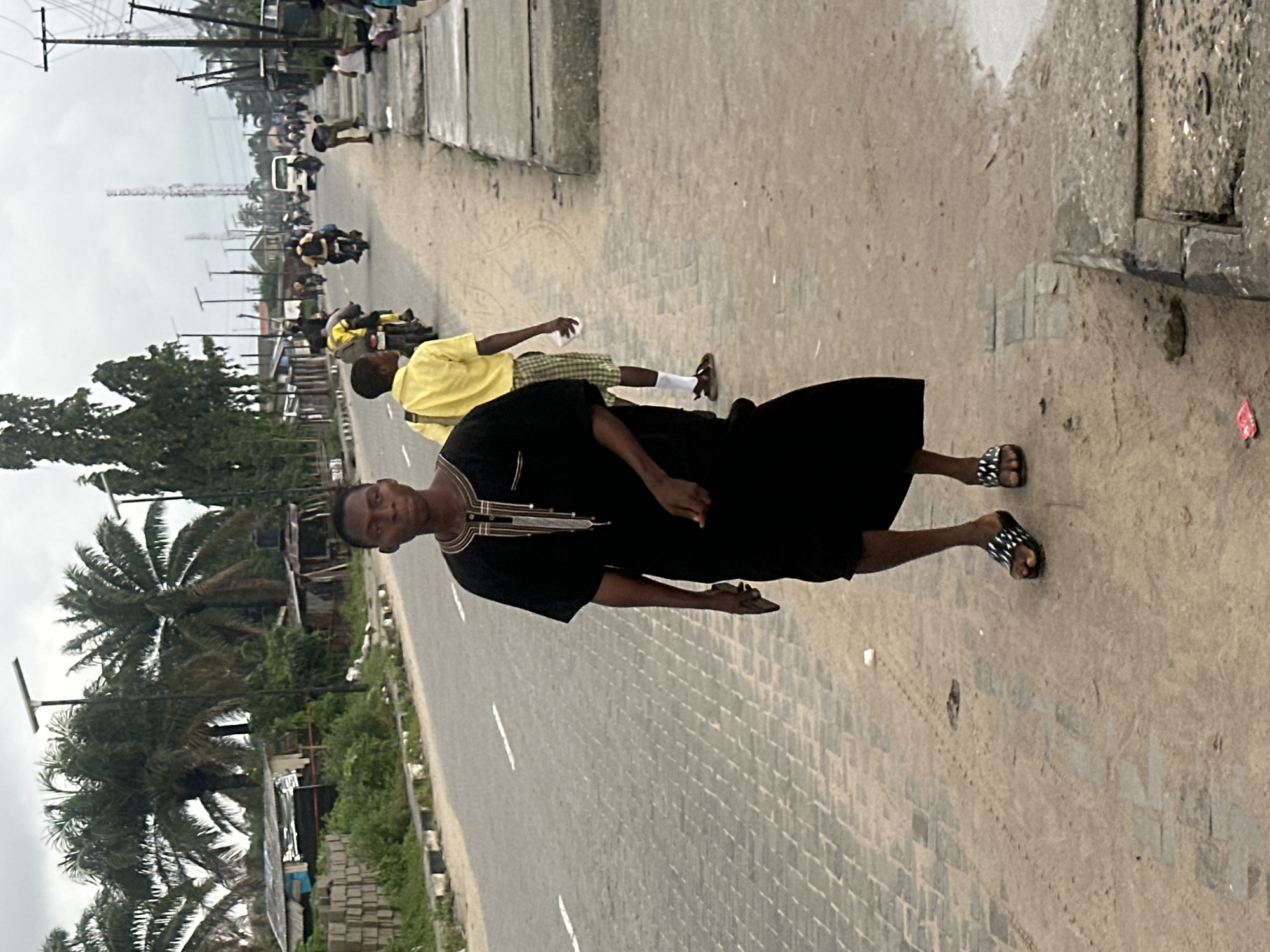 In a street scene in Badagry, Nigeria, a young Black man walks towards the viewer, holding a dark rectangular object in his right hand. He is wearing a black garment with gold and white embroidery around the neckline and down the front. His feet are adorned with black sandals with white geometric patterns. He is walking on a paved walkway beside a dusty road. In the background, several motorbikes and a white van are visible, along with other individuals. The environment includes palm trees, buildings, and overhead power lines. The sky is overcast.