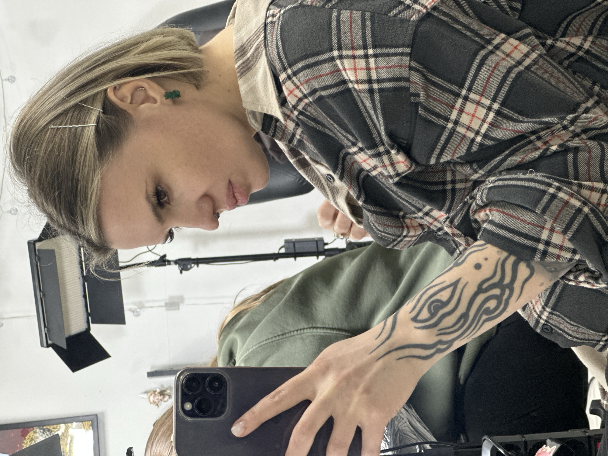An adult female is depicted taking a mirror selfie in an indoor setting within Rostov-on-Don, Russia, likely a studio or professional salon environment.
The primary subject, positioned in the right foreground, is a fair-skinned female with short, shoulder-length blonde hair, secured above her right ear with two silver bobby pins. She has a silver septum piercing and her left ear displays a small silver hoop piercing in the upper cartilage (helix) and a green gummy bear-shaped earring in the lobe. She is wearing a dark gray, beige, white, and red plaid collared shirt with a light beige collar. A black tribal-style tattoo featuring swirling lines and an abstract eye-like motif covers her left forearm. She holds a dark gray smartphone, identified as an iPhone Pro Max model, in her left hand, angling it to capture her reflection. Her fingernails are manicured with a light-colored polish.
A second individual is partially visible in the left background, seated and facing away from the camera. This individual is wearing a dark olive green hooded sweatshirt.
The background consists of a white wall. Professional studio lighting equipment is present, including a black LED panel light with barn doors mounted on a stand in the mid-background. A framed object with a dark border and ornate golden details is partially visible on the upper left wall. A small, white, gnome-like figurine is also affixed to the wall behind the secondary subject.