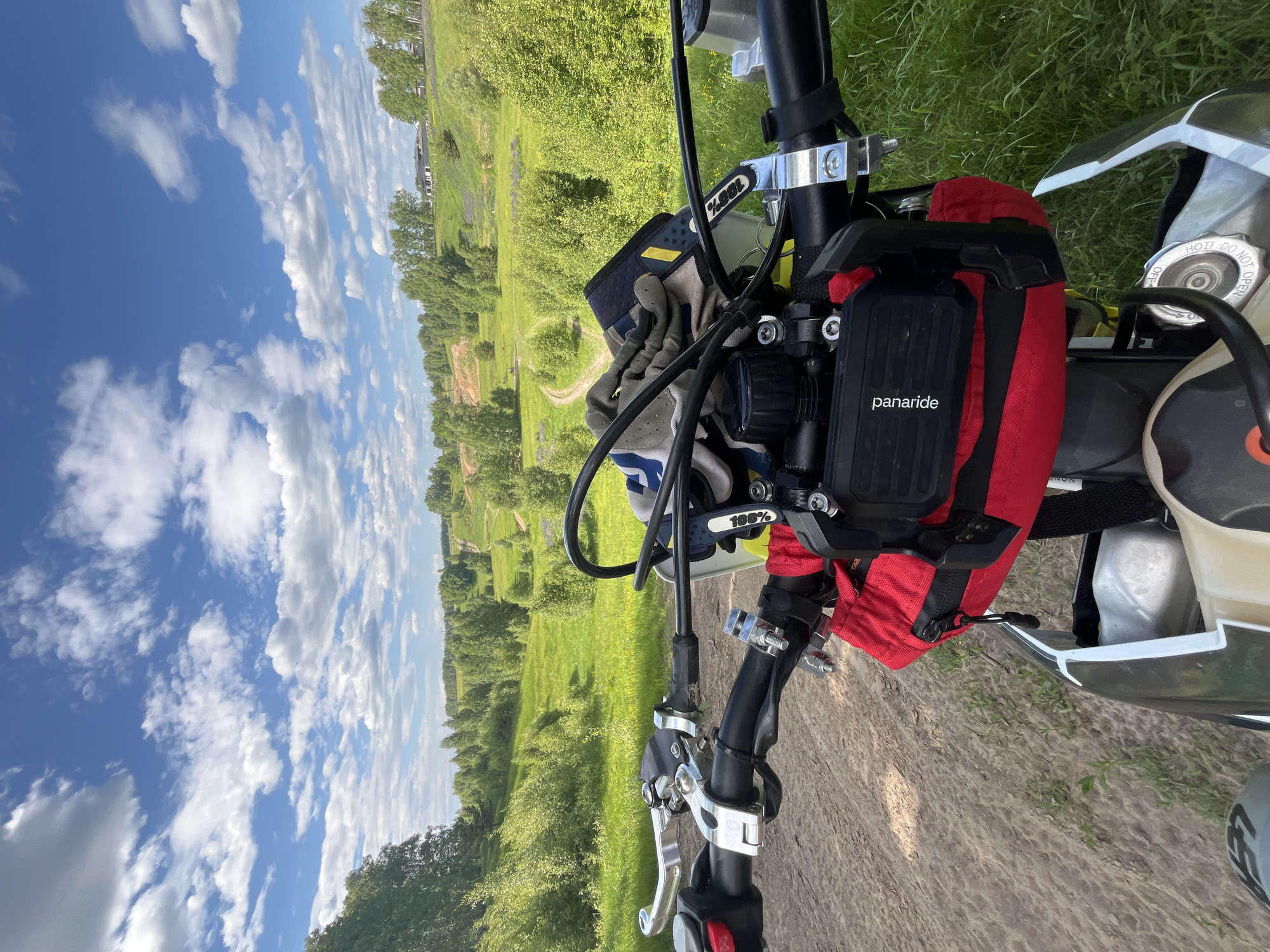 This is a first-person perspective shot from a motorcycle, stationary on a sandy dirt track, looking forward onto a rural landscape in Новочувашский, Russia. The scene depicts a bright, sunny day with a clear blue sky and numerous scattered white cumulus clouds.
In the immediate foreground, the handlebars of a dirt bike or enduro motorcycle are visible. The black handlebars feature silver clutch and brake lever assemblies on the left and right, respectively. A red button, possibly a kill switch, is on the left grip. A pair of grey, white, and blue motorcycle gloves, branded "100%", rests on the tank/handlebars. Centrally mounted on the handlebars is a black electronic device or holder, labeled "panaride", positioned above a red zippered tank bag or pouch. Portions of the motorcycle's white front fender, light-colored fuel tank, and black radiator shroud are also visible. The ground directly beneath the motorcycle is composed of light-brown sandy dirt with sparse green vegetation.
The midground features a winding dirt path or track that extends into the distance, flanked by lush green grass and numerous deciduous trees, suggesting a verdant, rolling terrain. This landscape consists of gentle hills and valleys covered in abundant greenery.
In the background, the rolling hills continue, densely covered with trees. On a distant hillside to the right, several modern houses or buildings with dark roofs are visible, indicating human settlement within the natural environment. The overall impression is one of a scenic rural area conducive to off-road motorcycling.
