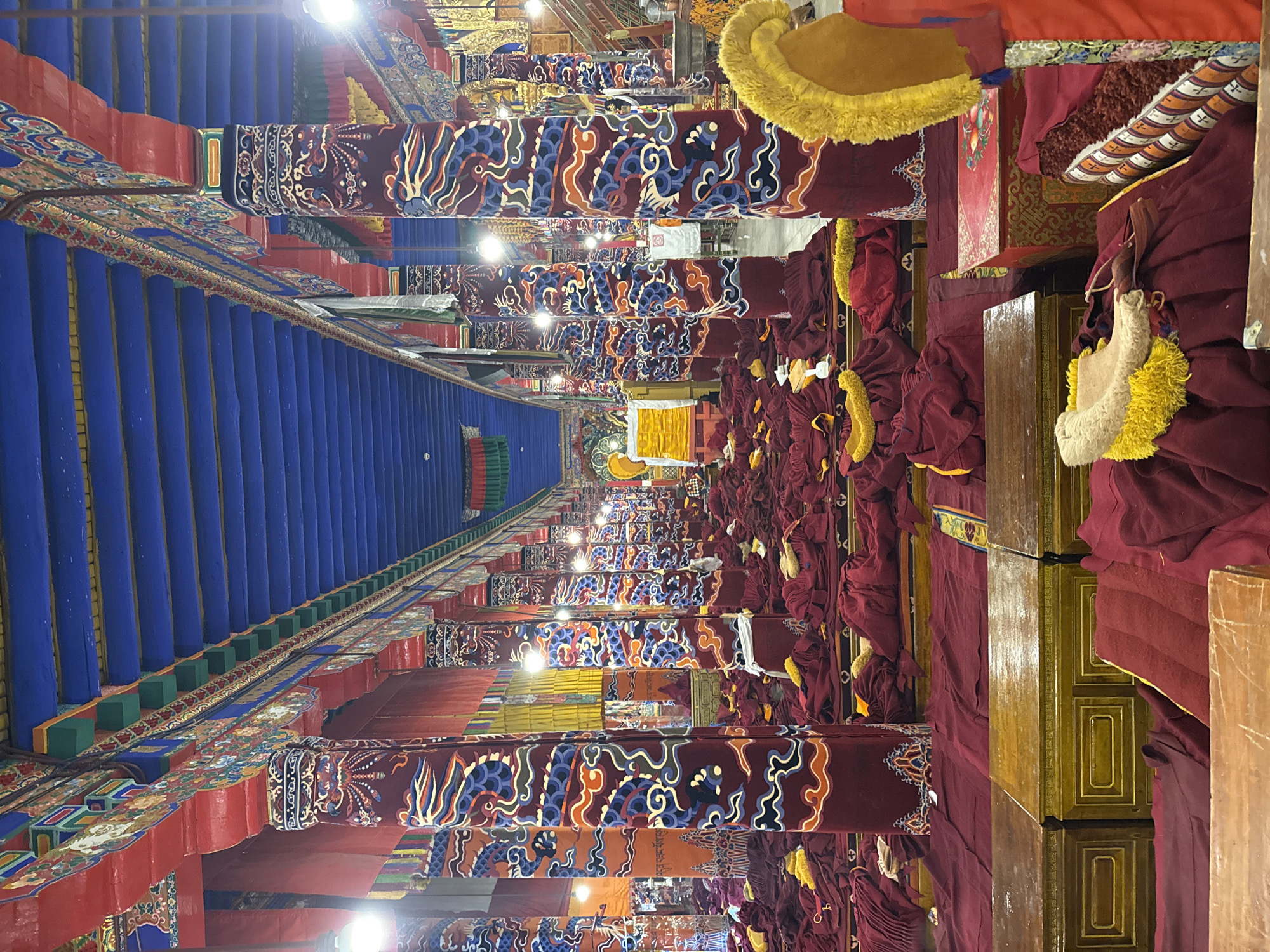 The image shows a hall in a Buddhist monastery, likely in Lhasa, China. Rows of monks in maroon robes are seated on cushions along both sides of a central aisle. They are wearing yellow head ornaments. The aisle floor is bright blue, bordered by ornate red and blue structures with decorative patterns. Pillars with similar patterns run along the sides of the hall. Overhead lighting illuminates the scene. The image is taken from an elevated perspective.