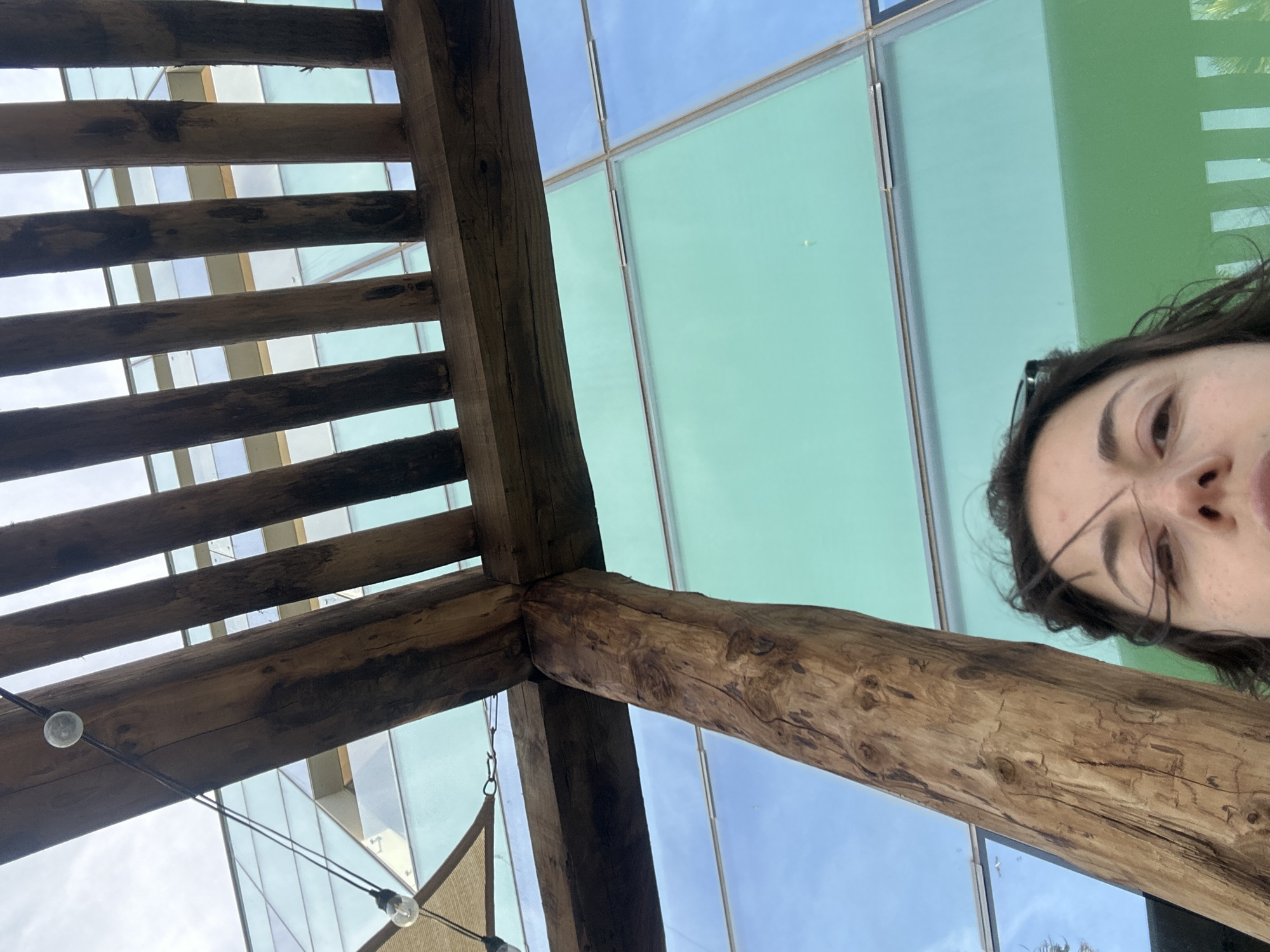 A low-angle shot features a woman's face partially visible on the right side of the frame, with the rest of the scene filled with architectural elements. Predominantly, there is an overlapping structure made of wooden beams, creating a grid-like pattern against a building exterior. The building's facade is composed of large panes of glass, some of which have a greenish tint, possibly reflective surfaces. The shot may be taken outdoors in Barcelona, Spain, as this is the only instruction.