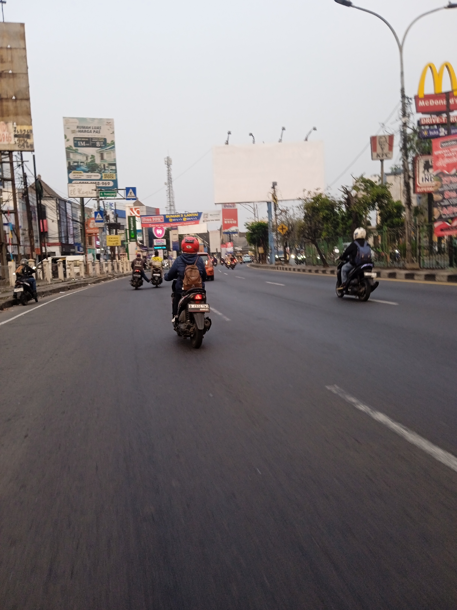The image shows a street scene in Bekasi, Indonesia. Several people are riding motorcycles on a multi-lane road. Billboards advertising various businesses, including McDonald's and KFC, are visible along the roadside. The sky is overcast, suggesting it might be late afternoon or early morning. The overall impression is of a busy urban environment.