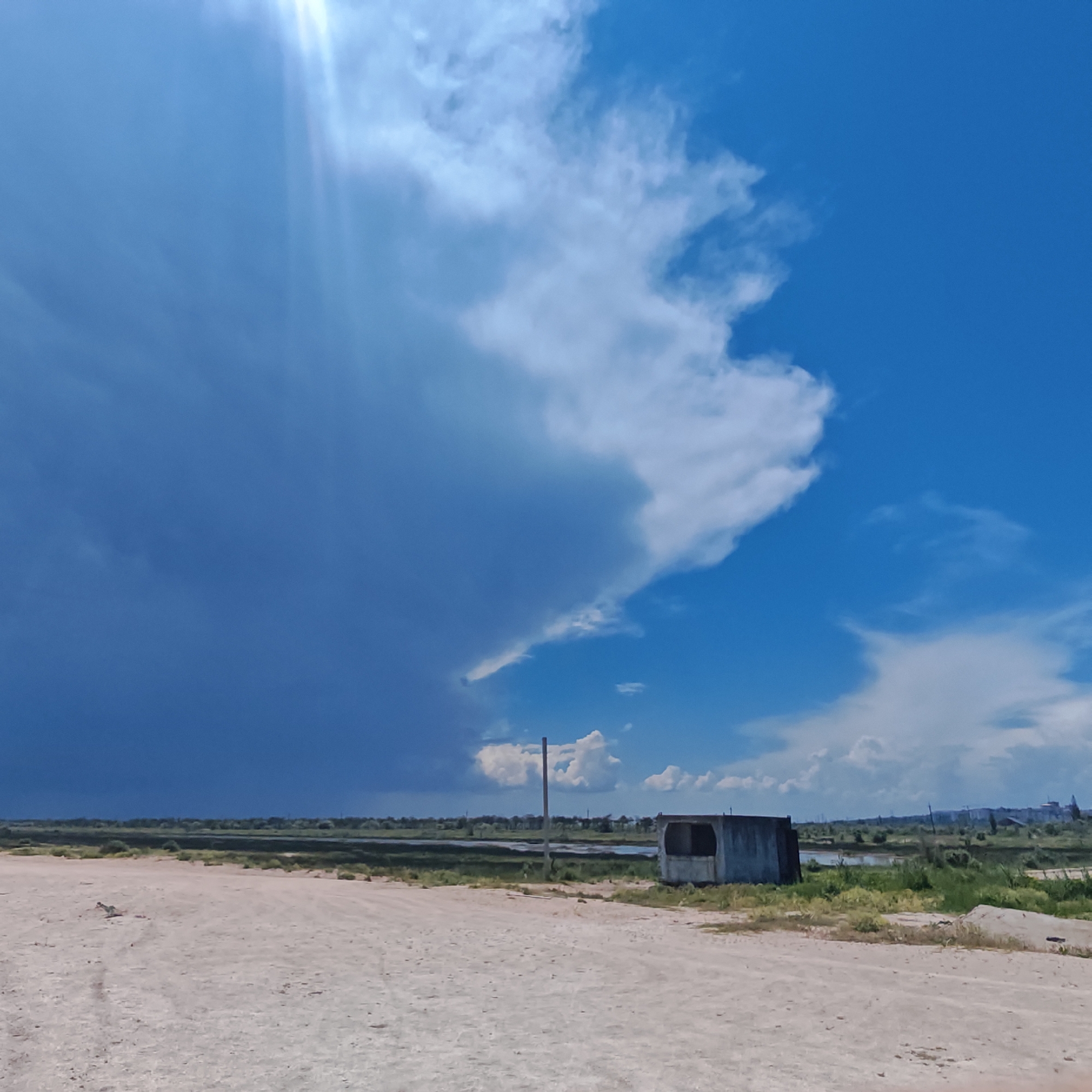 The image depicts a broad, sandy foreground leading to a flat landscape with sparse vegetation and a body of water in the mid-ground. The sky is a prominent feature, with a dramatic contrast between a dark, ominous storm cloud on the left and a bright, clear blue sky with scattered white clouds on the right. A single, weathered concrete structure, possibly a small shed or shelter, sits in the mid-ground to the right. A wooden pole, likely a utility pole, is also visible. In the far distance, a hazy skyline suggests a populated area. The overall scene conveys a sense of an impending storm or the aftermath of one.